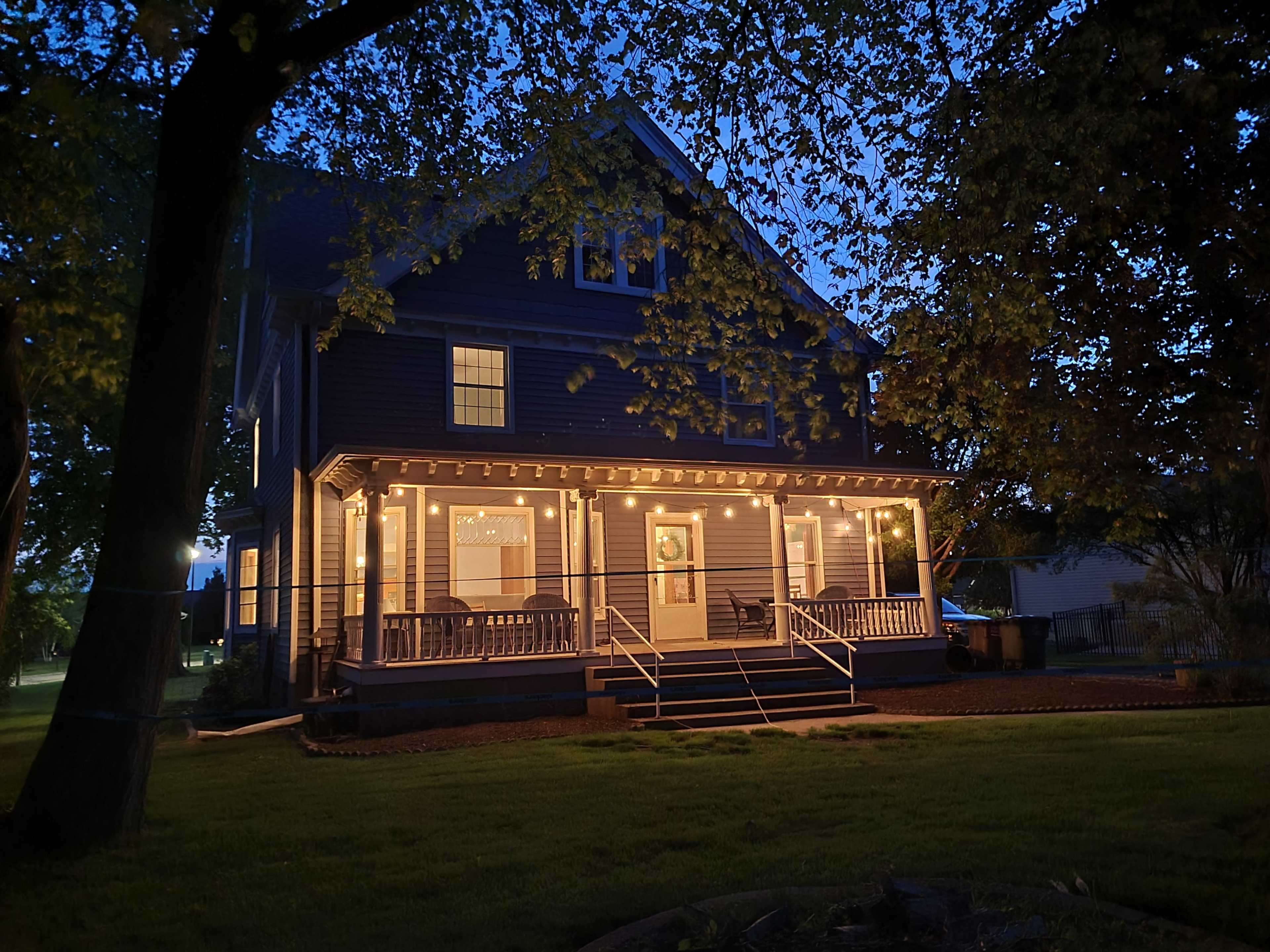 A two-story house with festive string lights illuminating the porch is surrounded by trees at dusk.