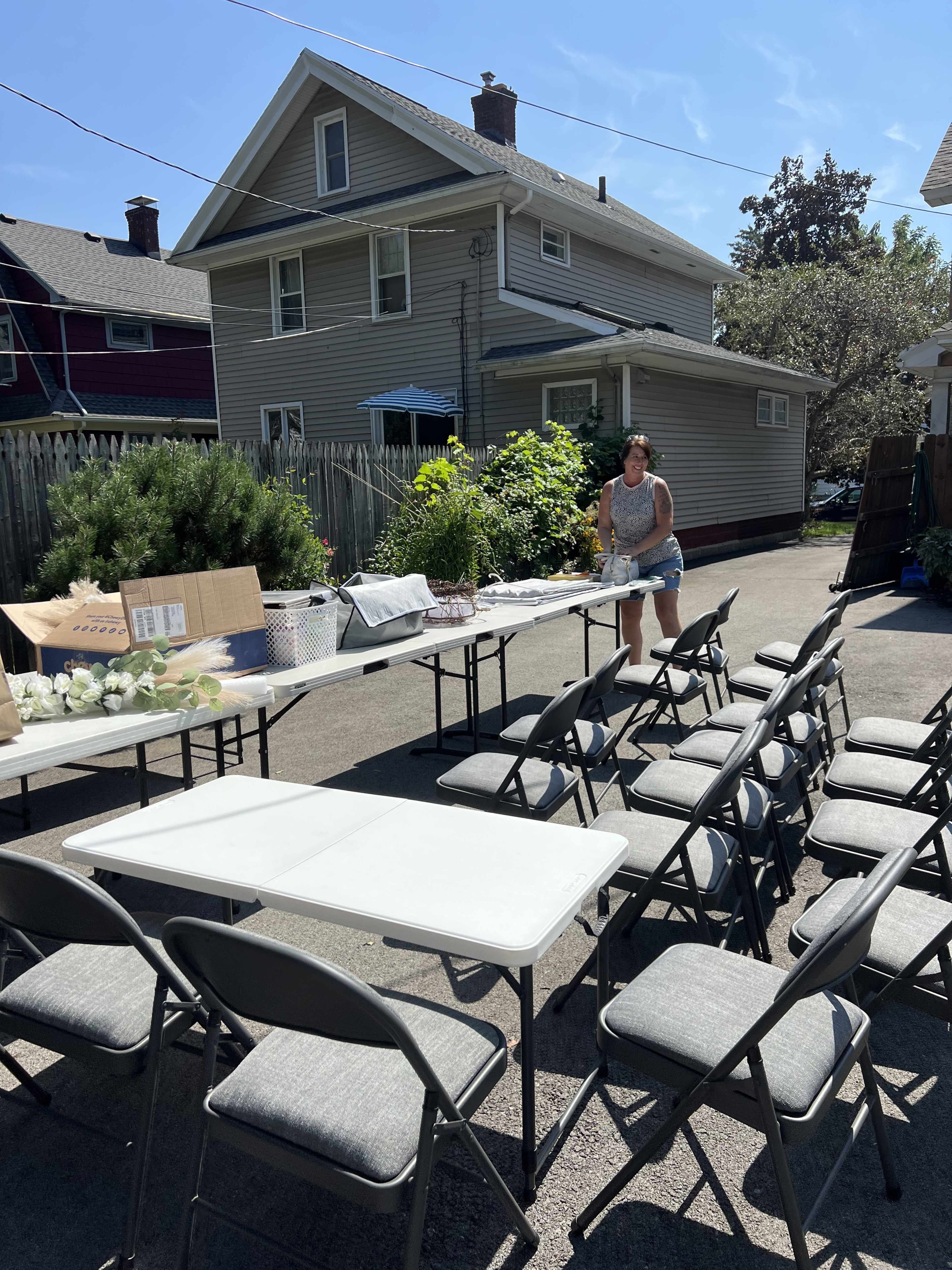 A woman arranges items on a long table surrounded by folding chairs in a sunny backyard near a house.