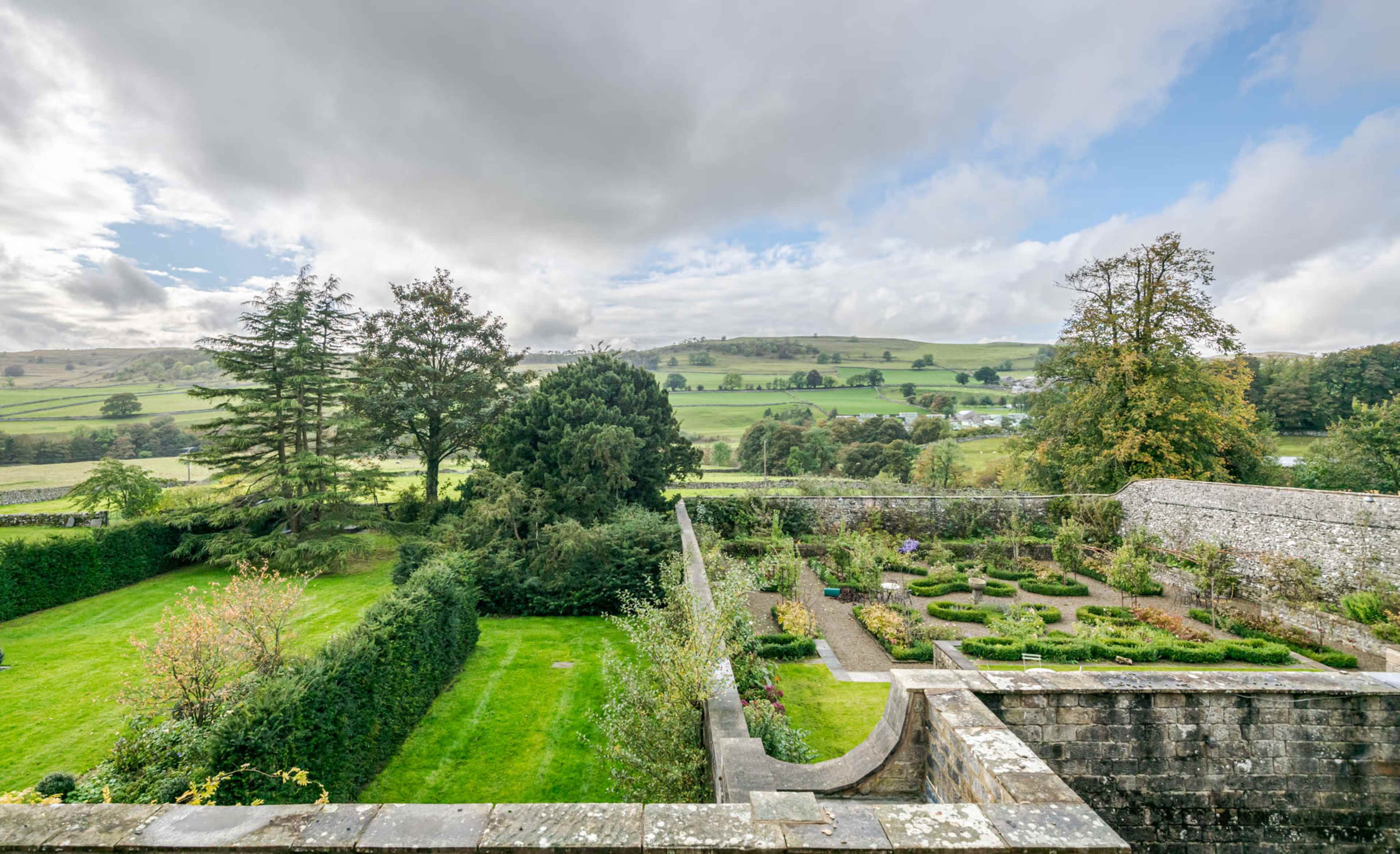 A lush green landscape with neatly arranged garden beds surrounded by stone walls, under a partly cloudy sky.