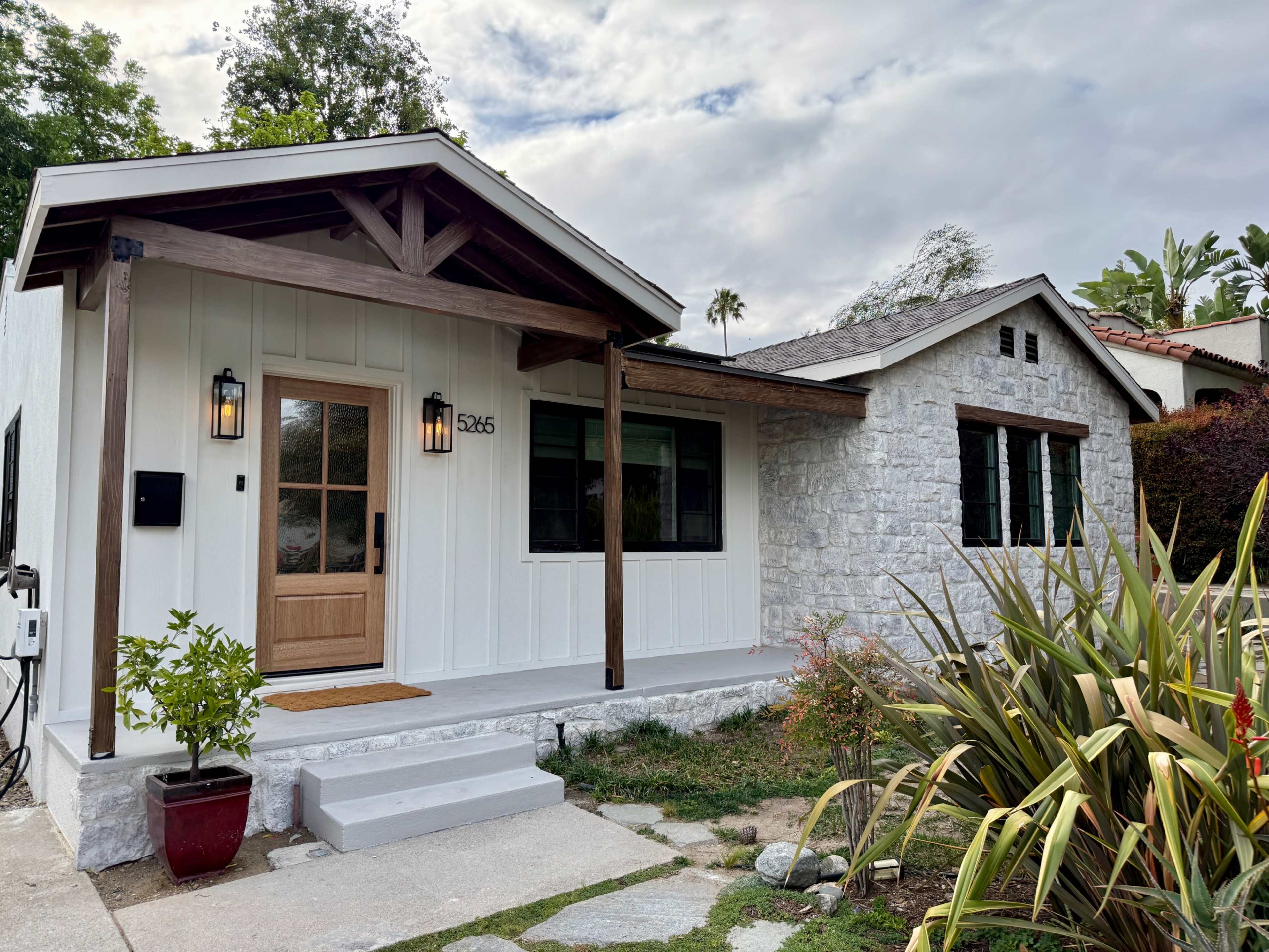 The image shows a modern single-story house featuring a wooden front door, a porch area, and a combination of white siding and stone exterior.