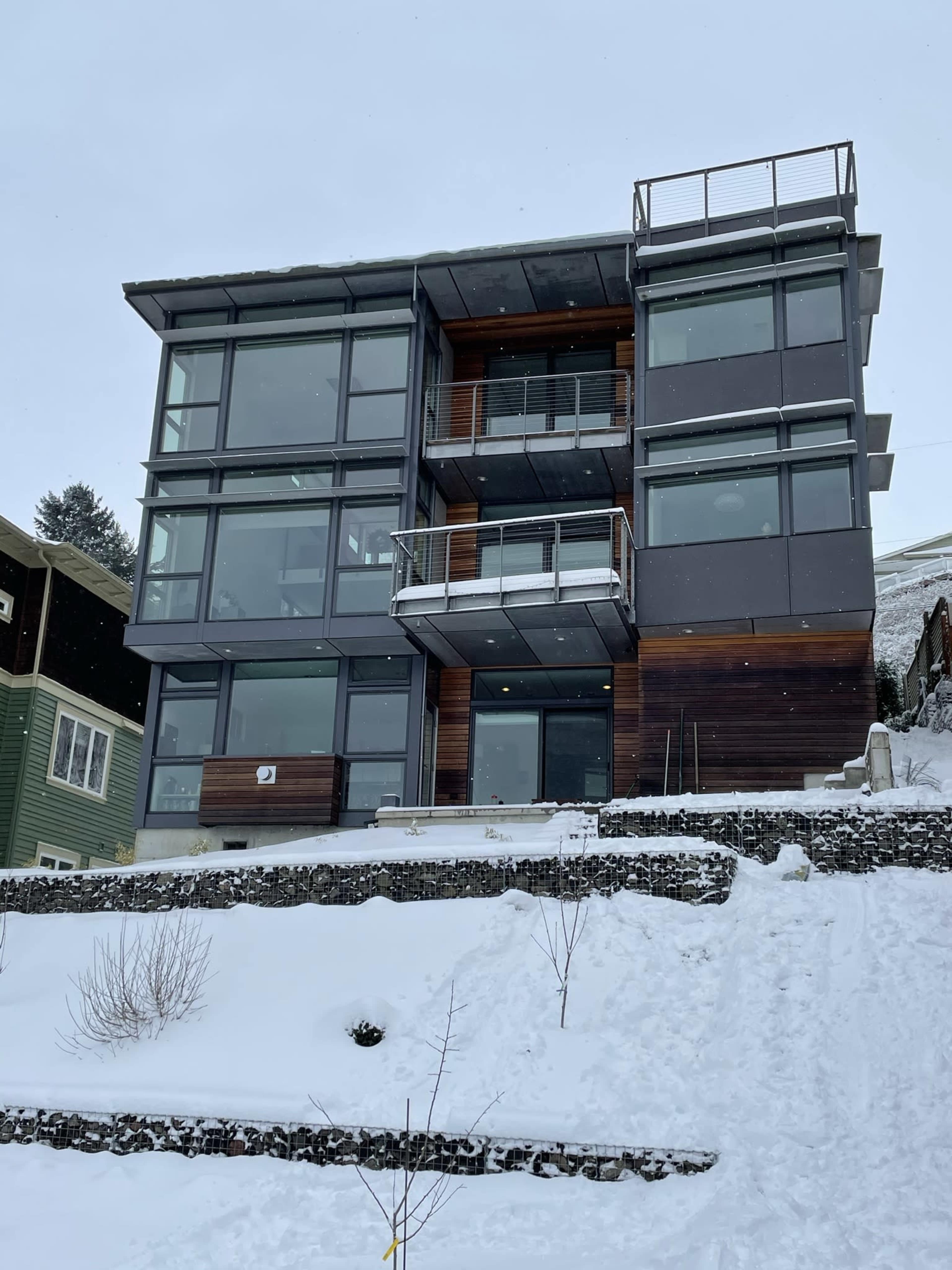 A modern, multi-story house with large glass windows and wooden accents is surrounded by snow and slopes.