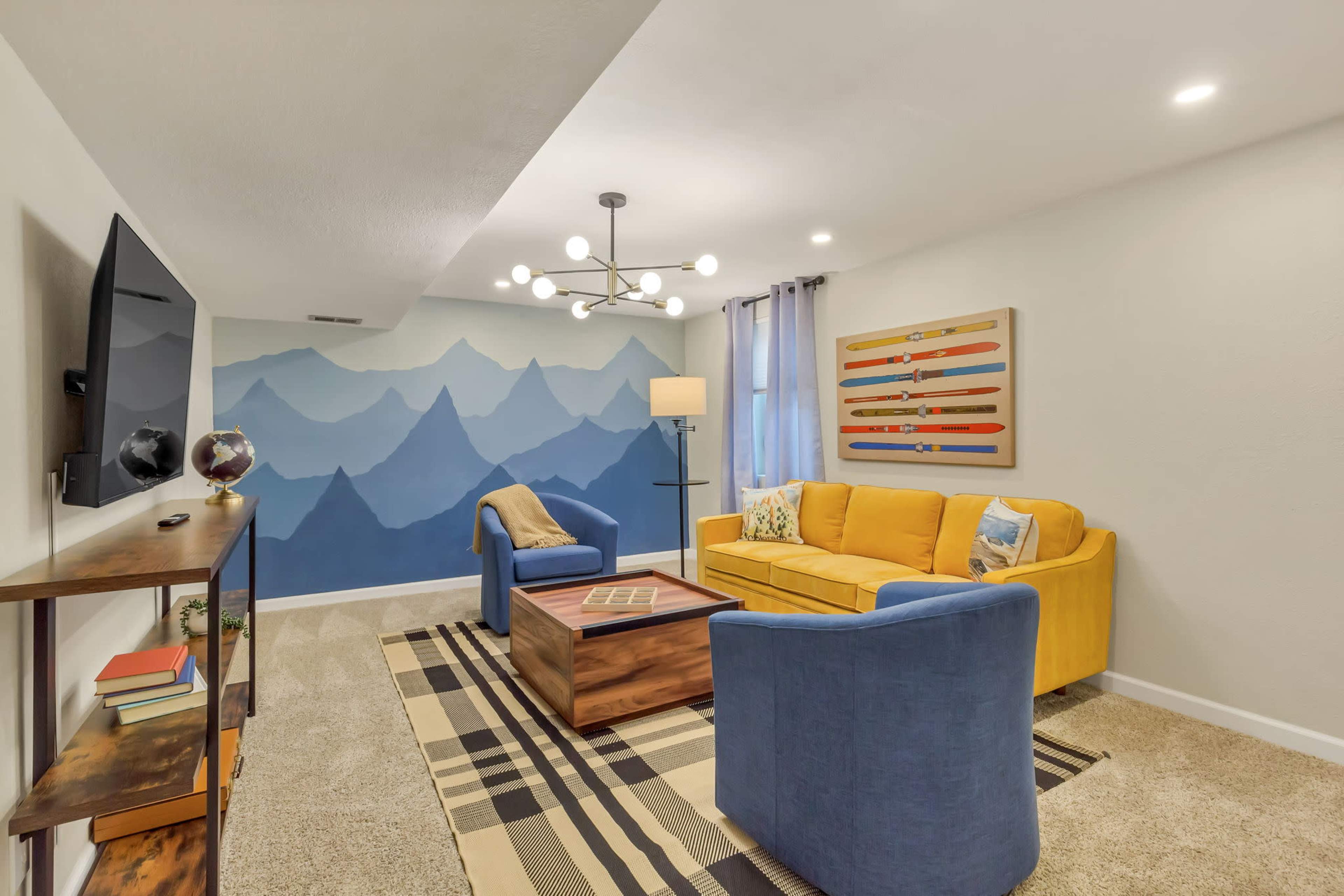 The living room features a yellow couch, blue chairs, a wooden coffee table, and a wall mural depicting mountain silhouettes.