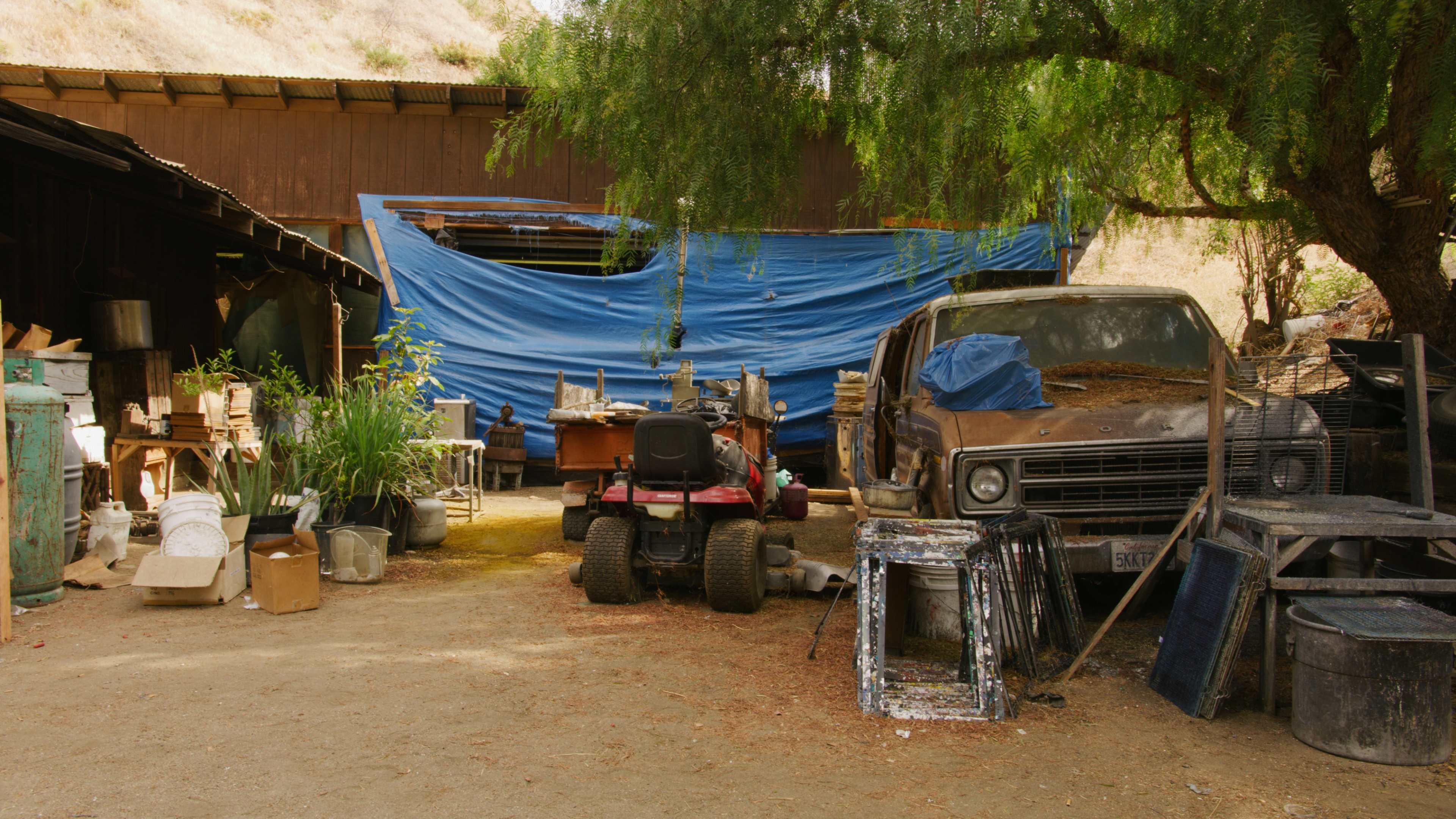 The scene shows a cluttered outdoor area with various tools, an old truck, and a blue tarp covering a structure.