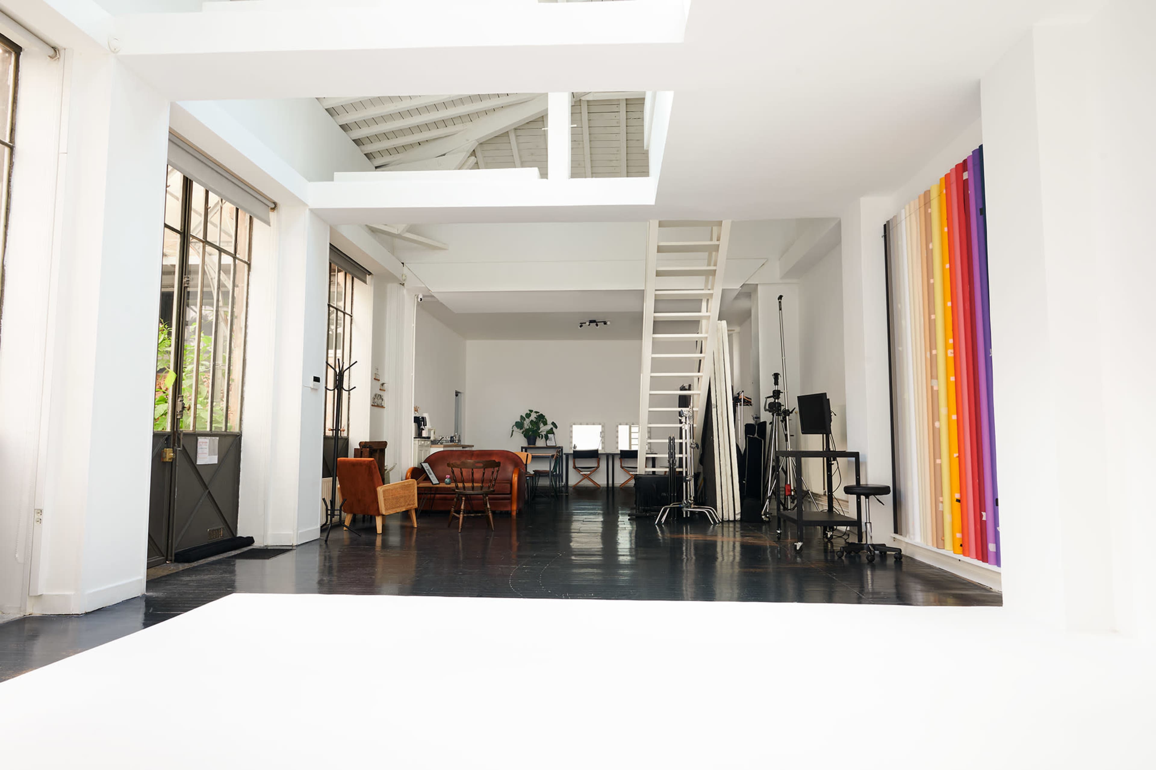 A spacious, bright studio with a wooden ladder, furniture including a brown couch, and large windows letting in natural light.
