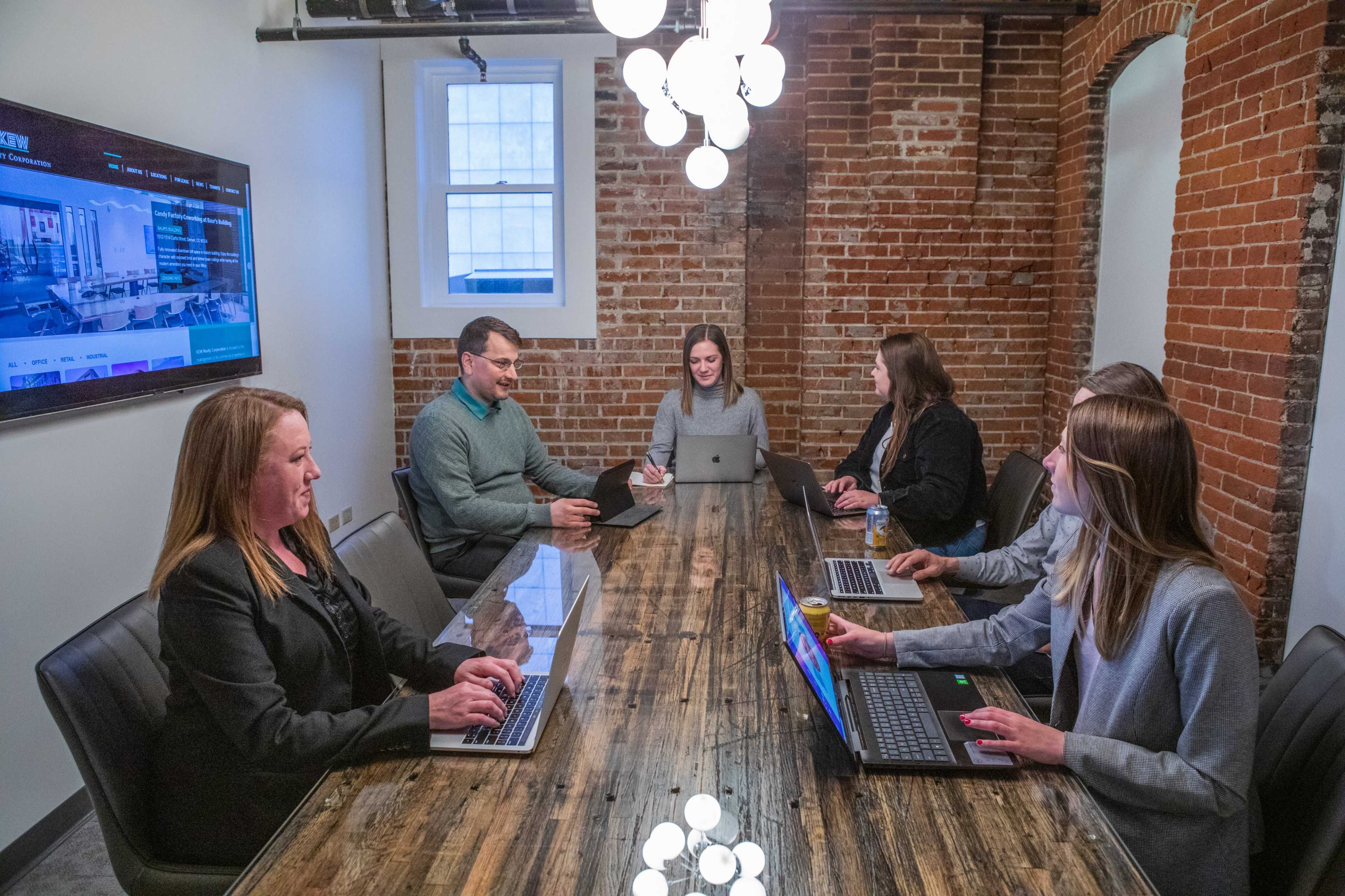 A group of six people sit around a wooden conference table in a meeting room with exposed brick walls, each using laptops and engaged in conversation.