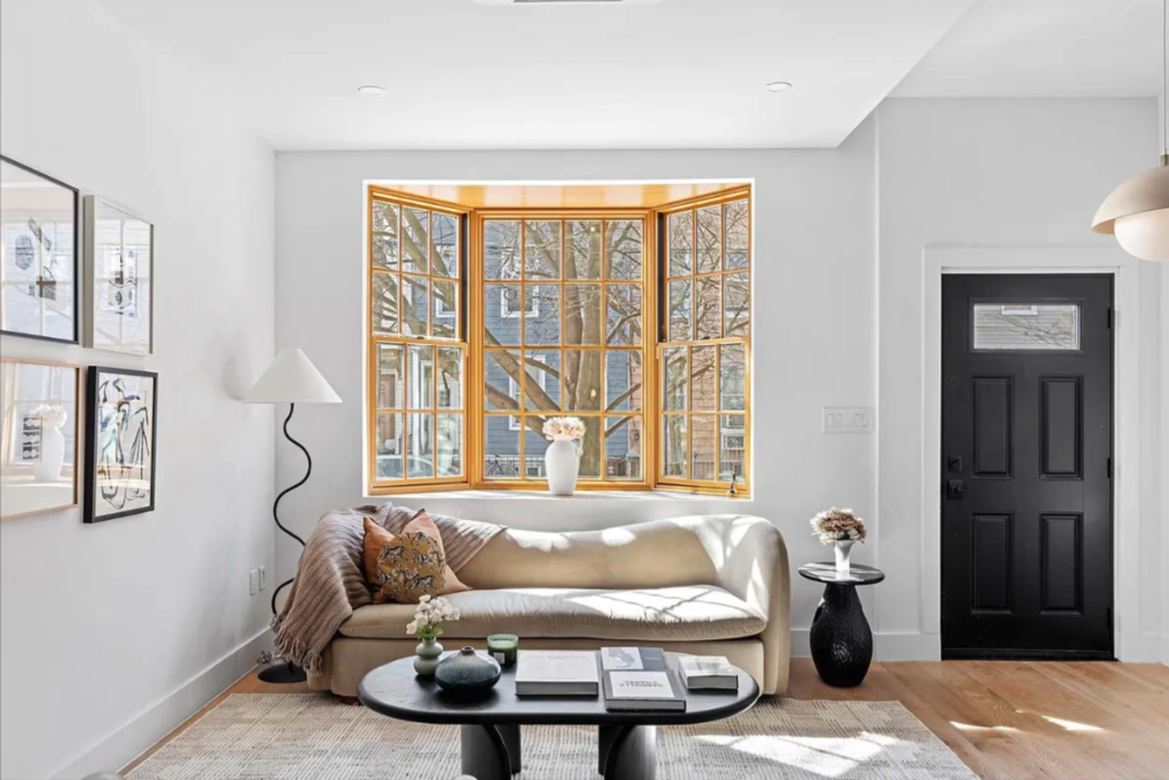 A bright living room features a large bay window, a cream-colored sofa, a round black coffee table, and minimalist wall decor.