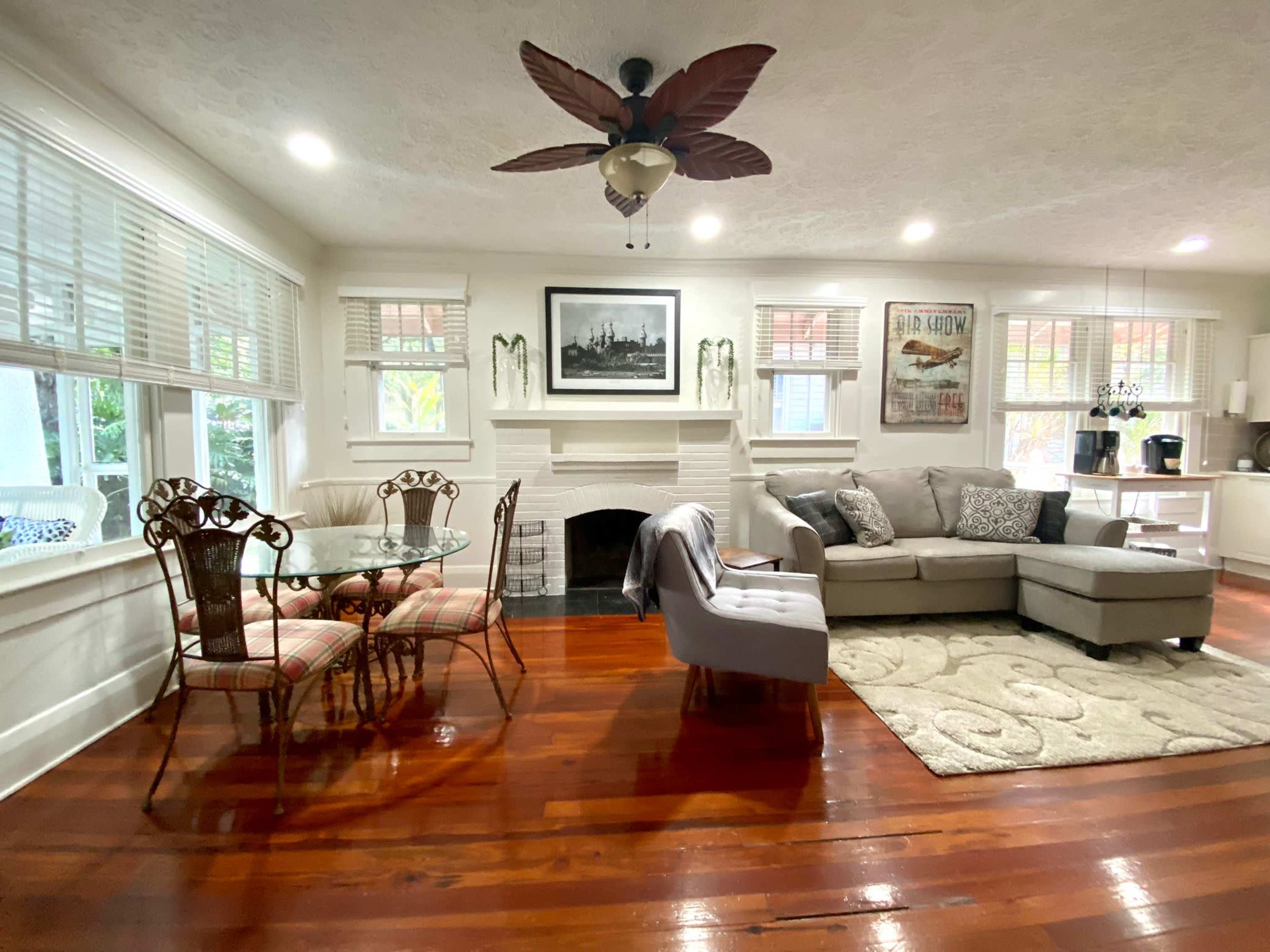 The image shows a living room with a sofa, a round dining table, a fireplace, and large windows, all set against hardwood flooring.