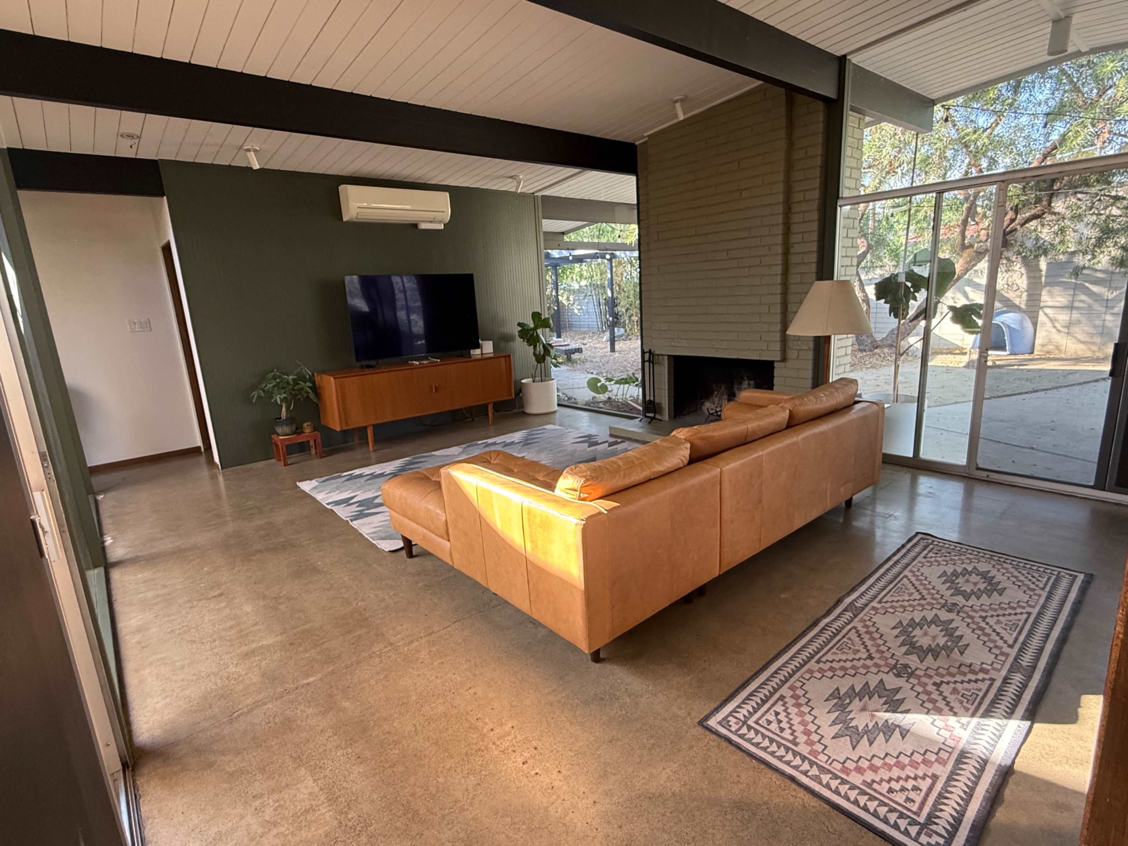 The image shows a living room featuring a brown leather sectional sofa, a television mounted on the wall, and a large window overlooking an outdoor space with trees.