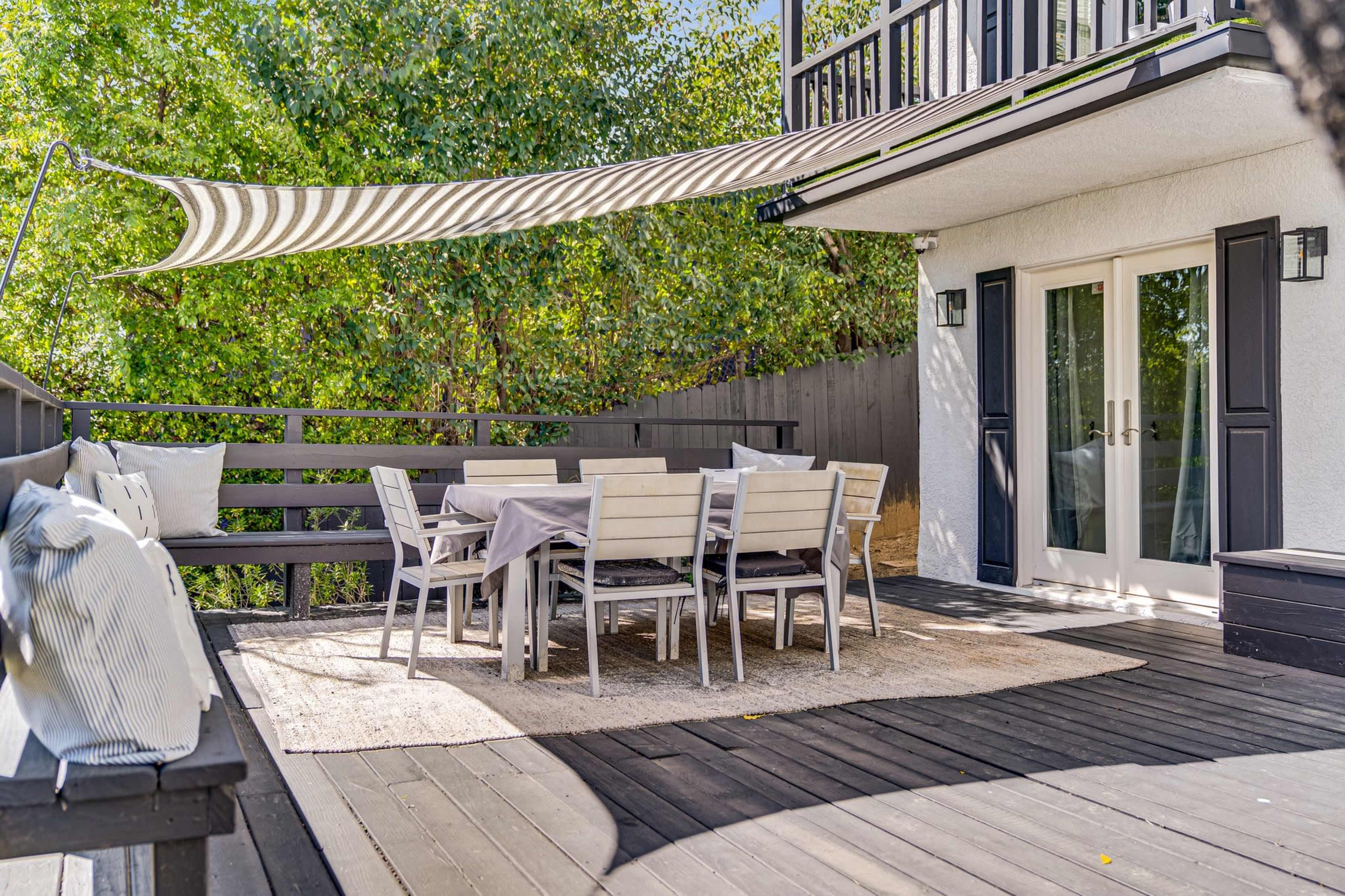 A spacious wooden deck features a dining table surrounded by chairs, with a shaded area provided by a striped awning.