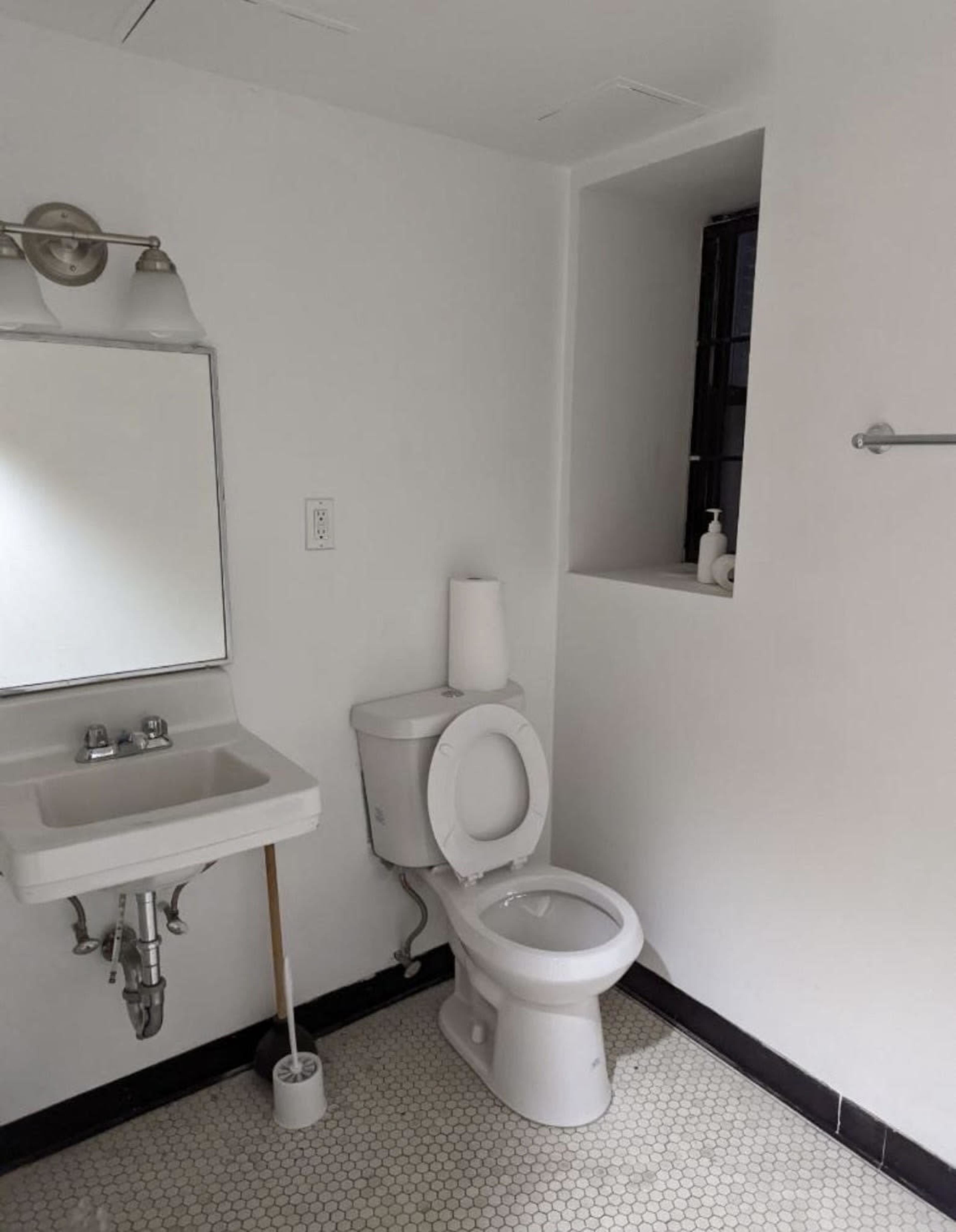 A bathroom featuring a toilet next to a small sink and a mirror, with a window visible in the background.