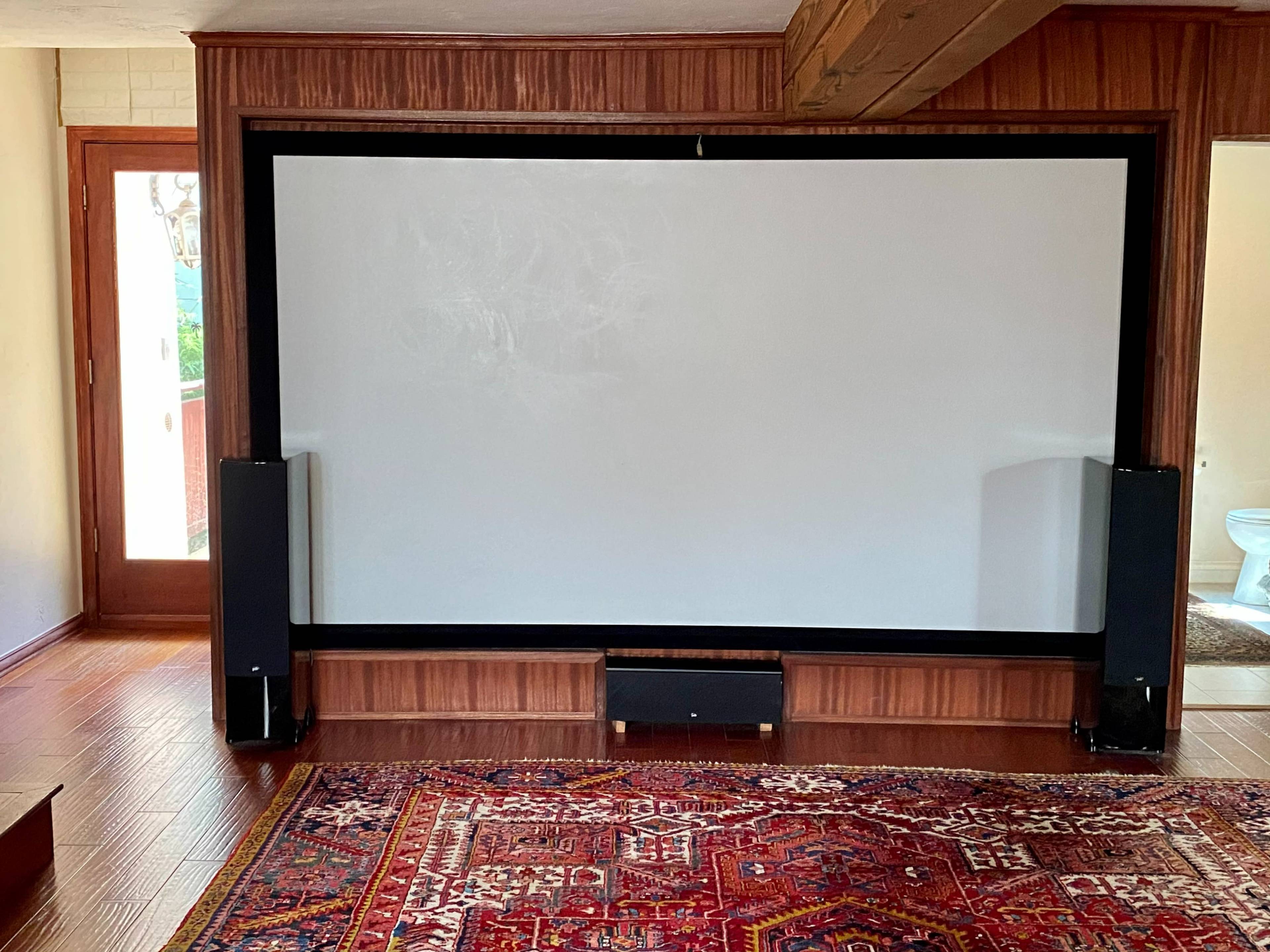 A home theater setup with a large projection screen framed by two black speakers, situated in a room with wooden paneling and a patterned rug.