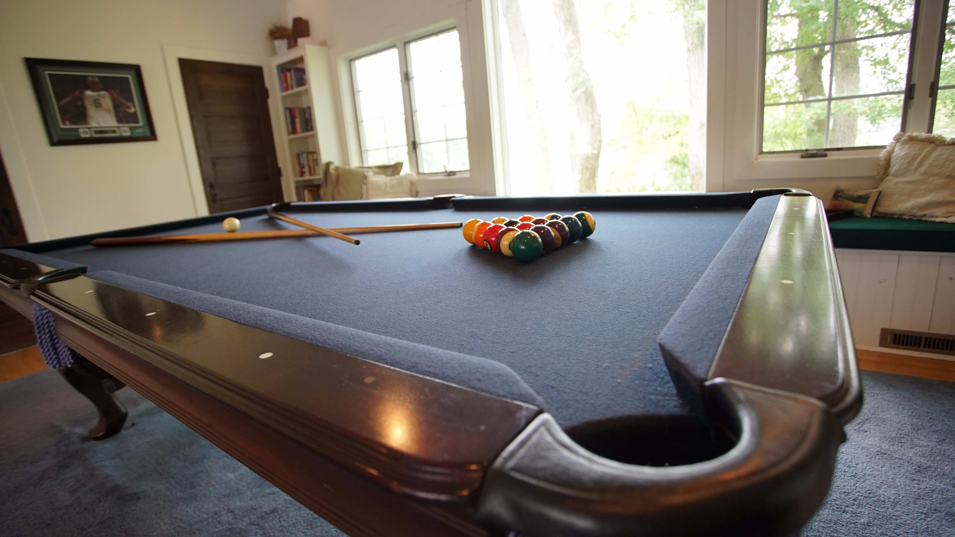 A pool table with a complete set of balls arranged in a triangle is positioned in a well-lit room featuring large windows.