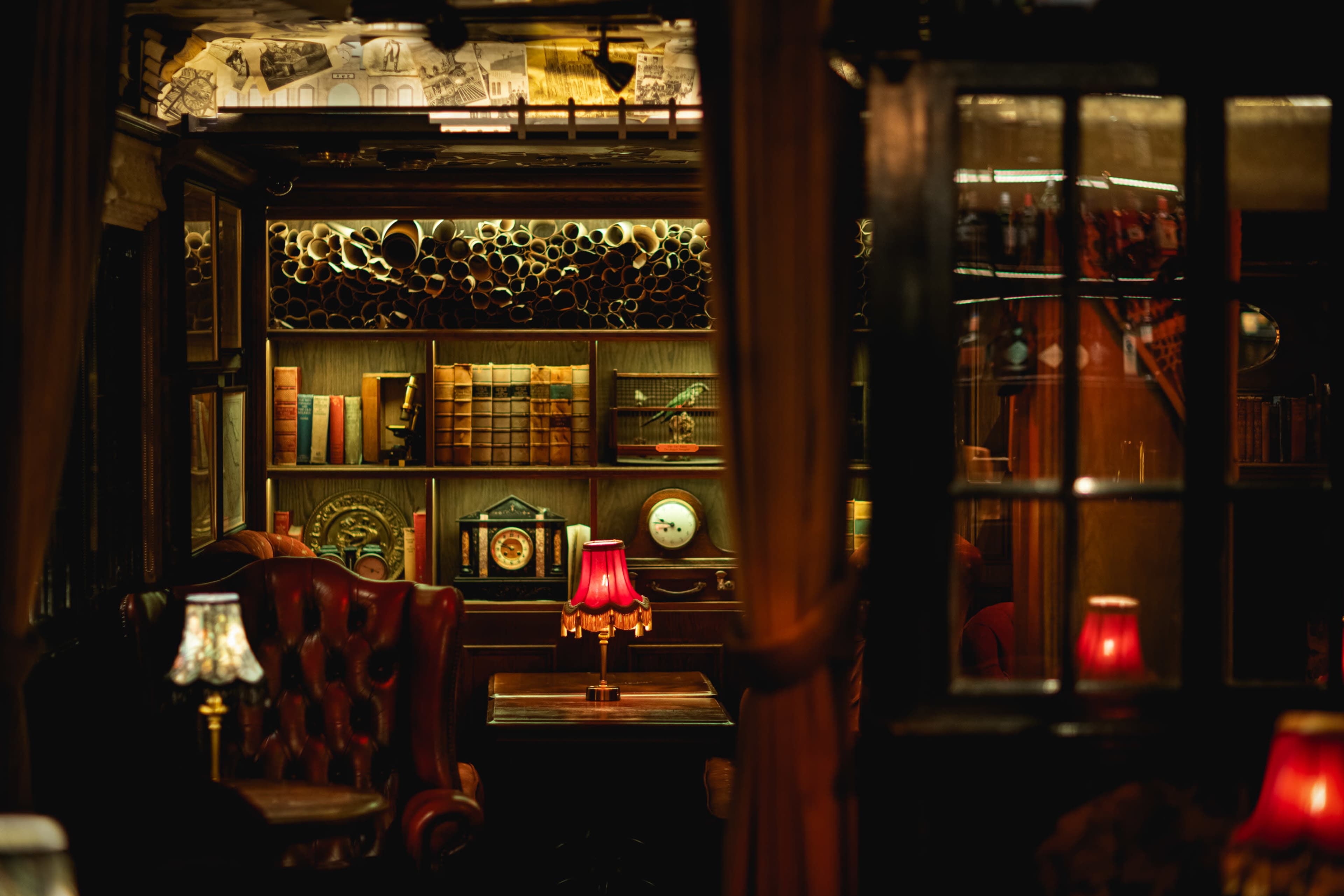A cozy interior of a dimly lit pub features leather armchairs, a wooden table, and shelves filled with books and antique items, illuminated by table lamps.