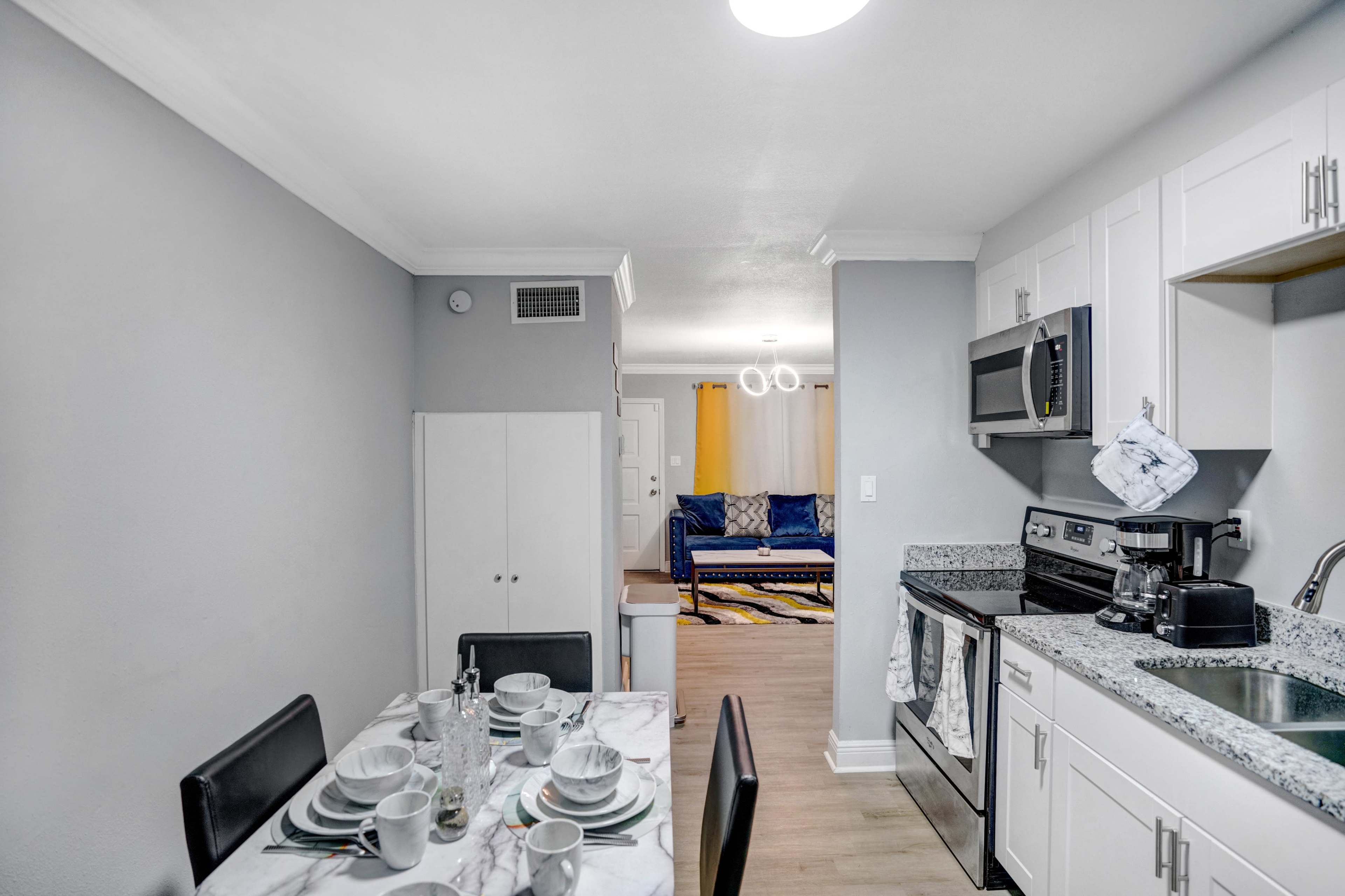 The image shows a modern kitchen area with a dining table set for four, adjacent to a living room featuring a blue sofa and light-colored decor.