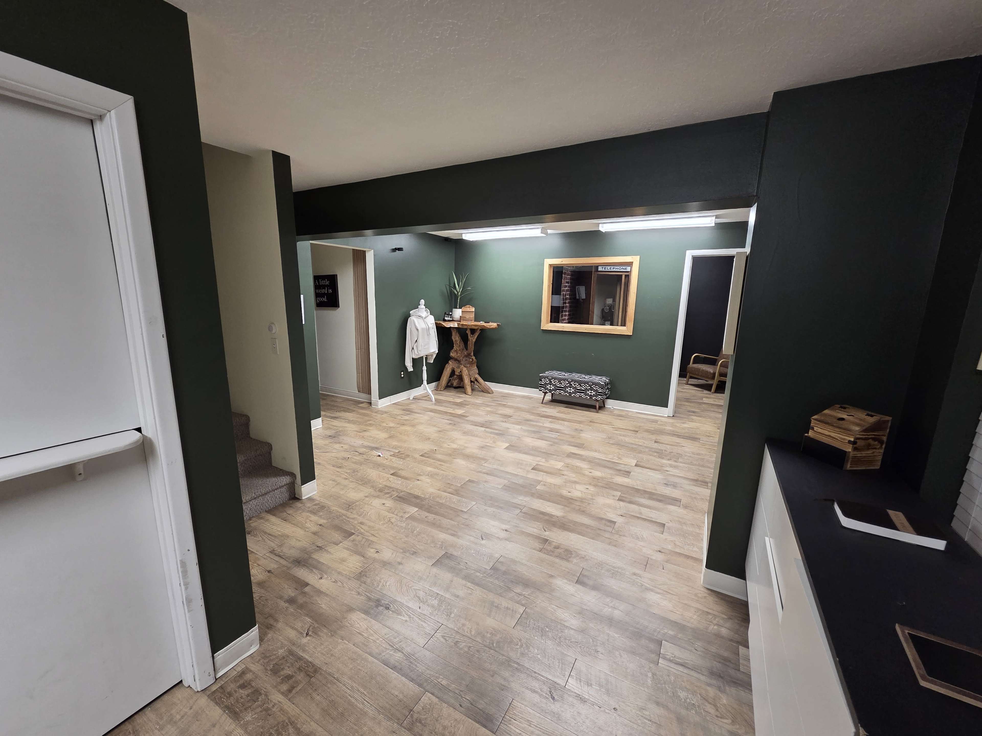 The image shows a spacious, well-lit room with wooden flooring, green walls, a staircase, and furniture including a round wooden table and a decorative bench.