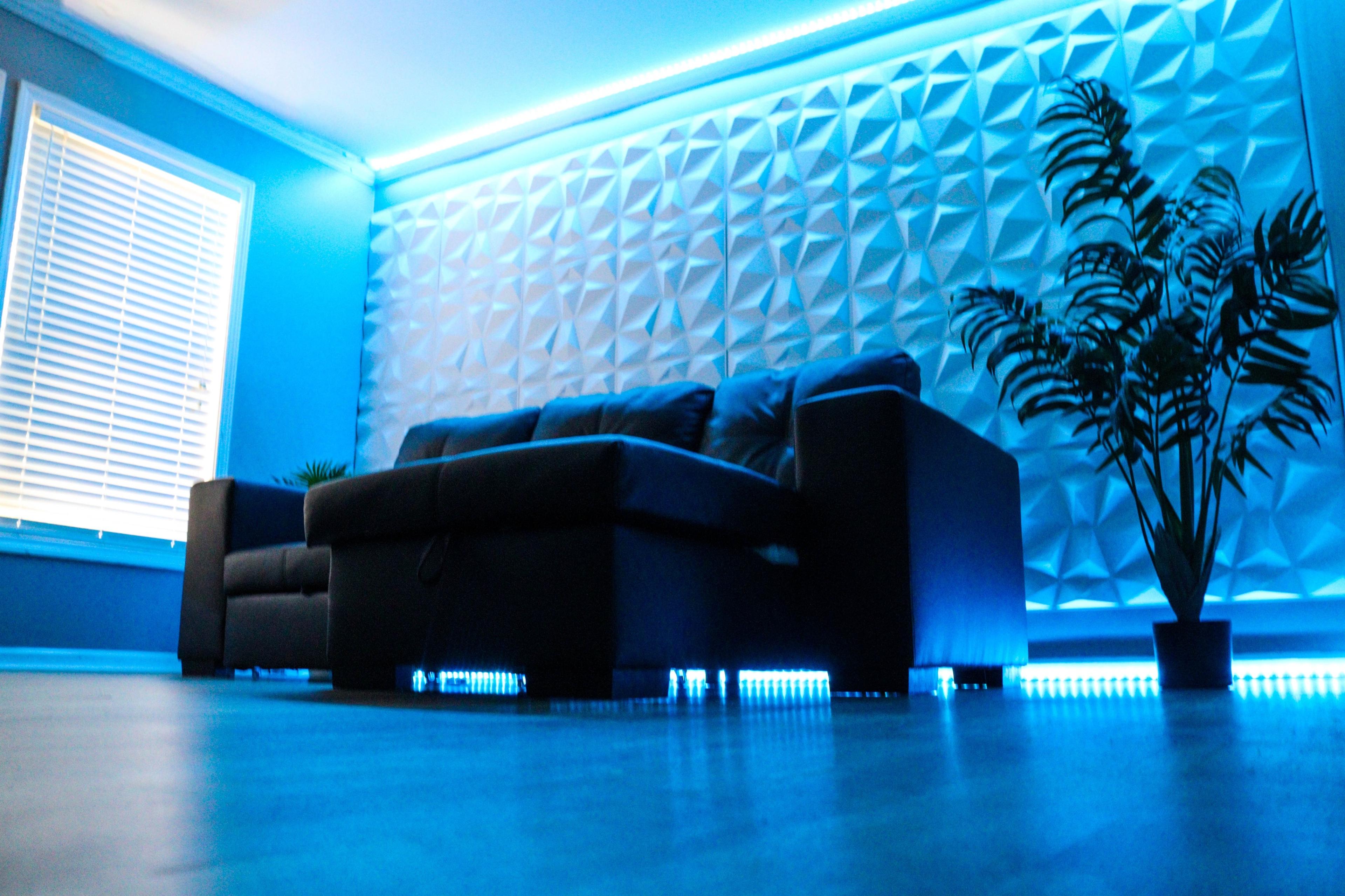 A modern living room features a black sectional sofa against a textured white wall, illuminated by blue LED lights.