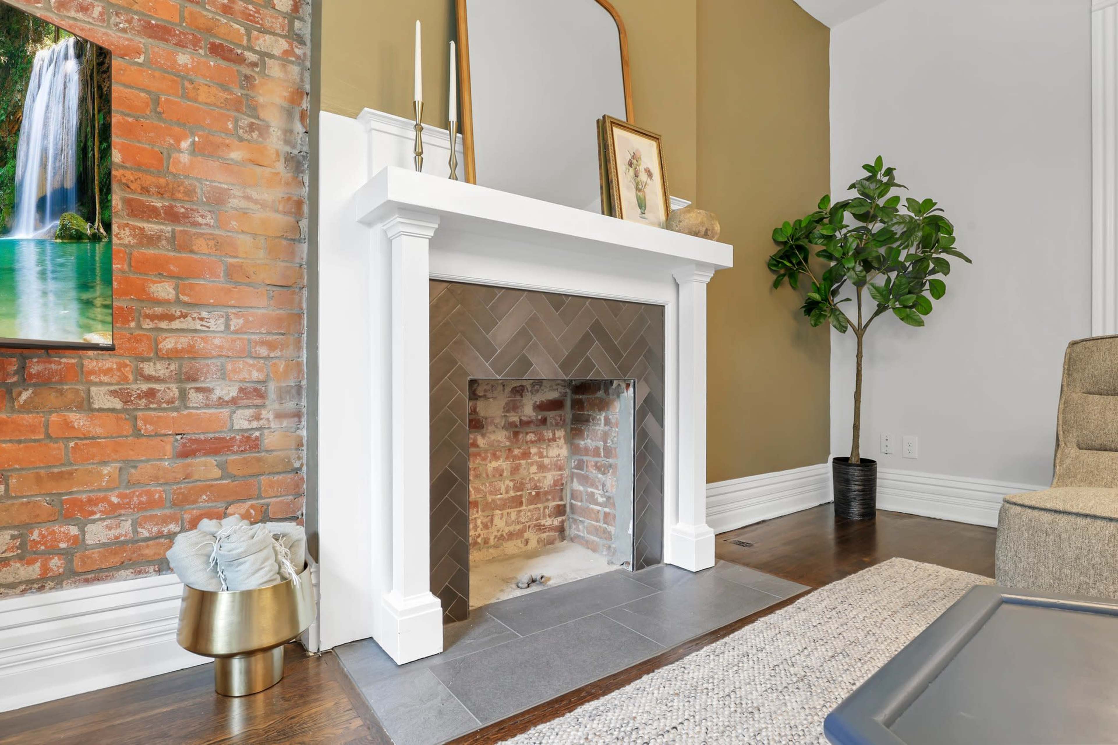 A modern living room with a white fireplace adorned with a herringbone pattern and surrounded by exposed brick walls, complemented by a mirror and a decorative plant nearby.