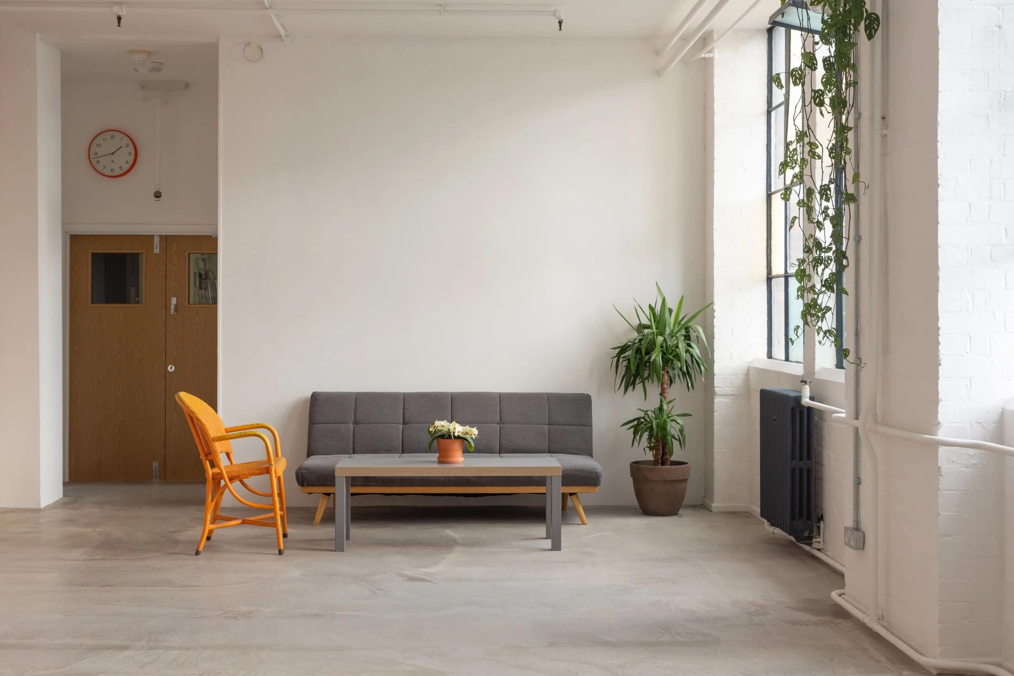 A gray sofa and a wooden table are positioned in a spacious room with large windows, a potted plant, and an orange chair.