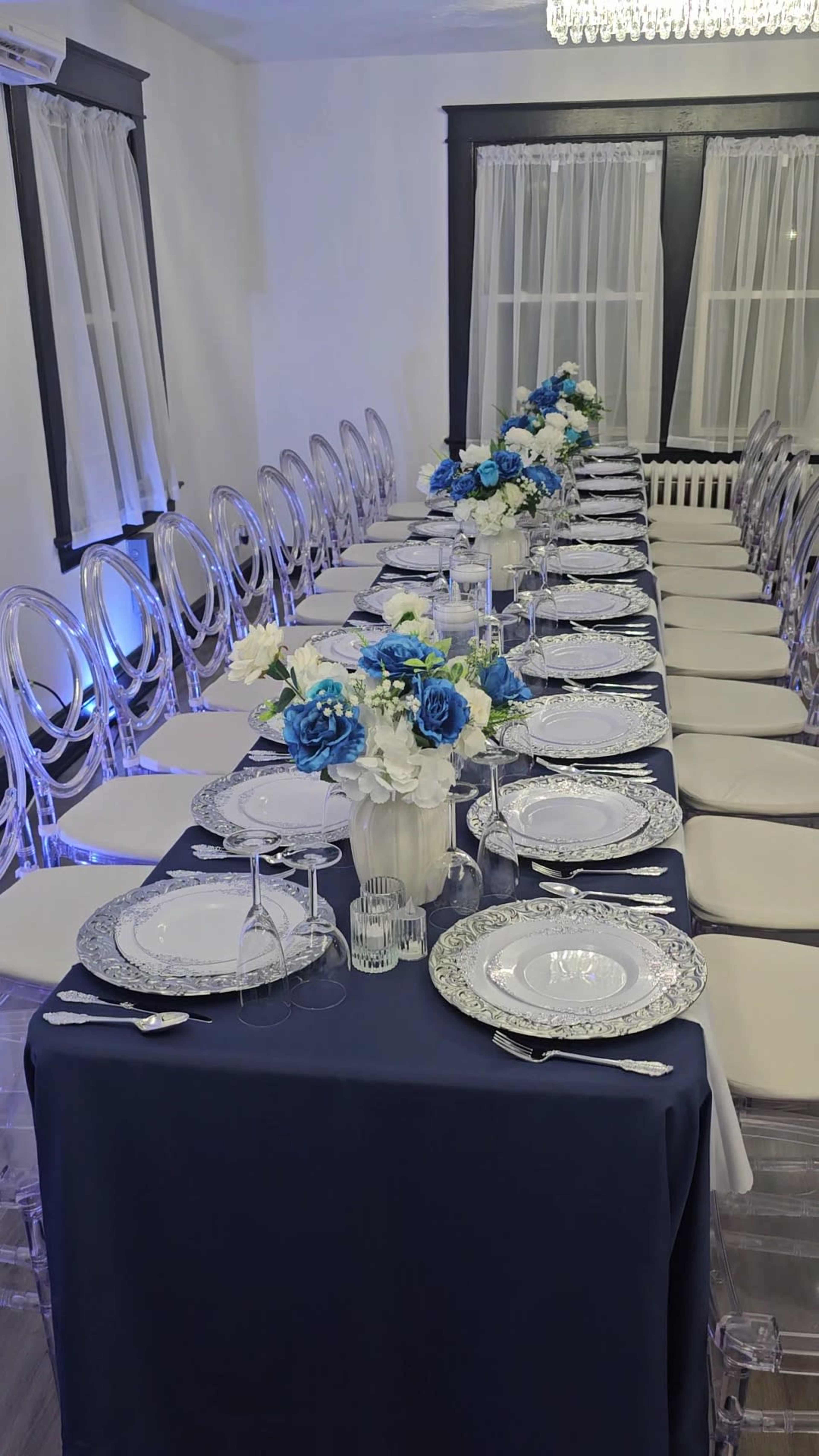 A long banquet table is set with white and blue floral arrangements, elegant plates, and glassware, surrounded by clear chairs in a well-lit room.