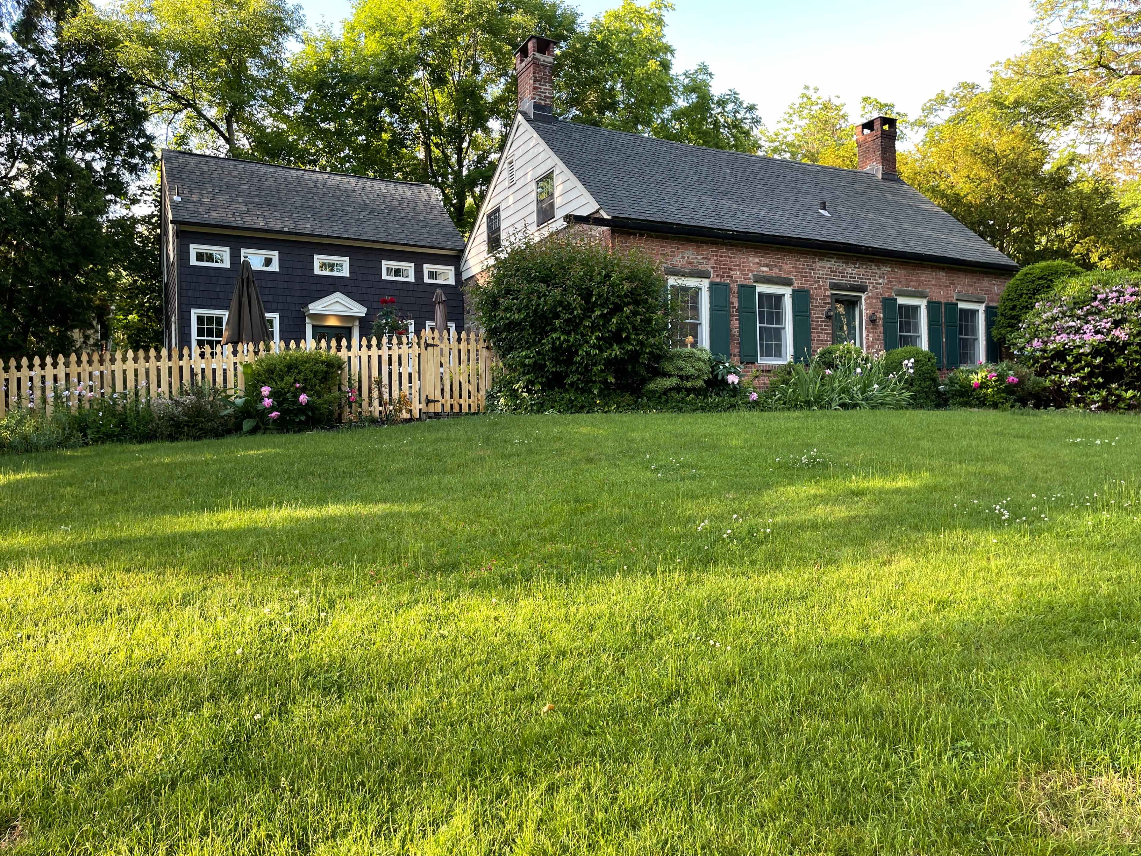 A brick house with green shutters is situated next to a gray building, surrounded by a lush lawn and a white picket fence.