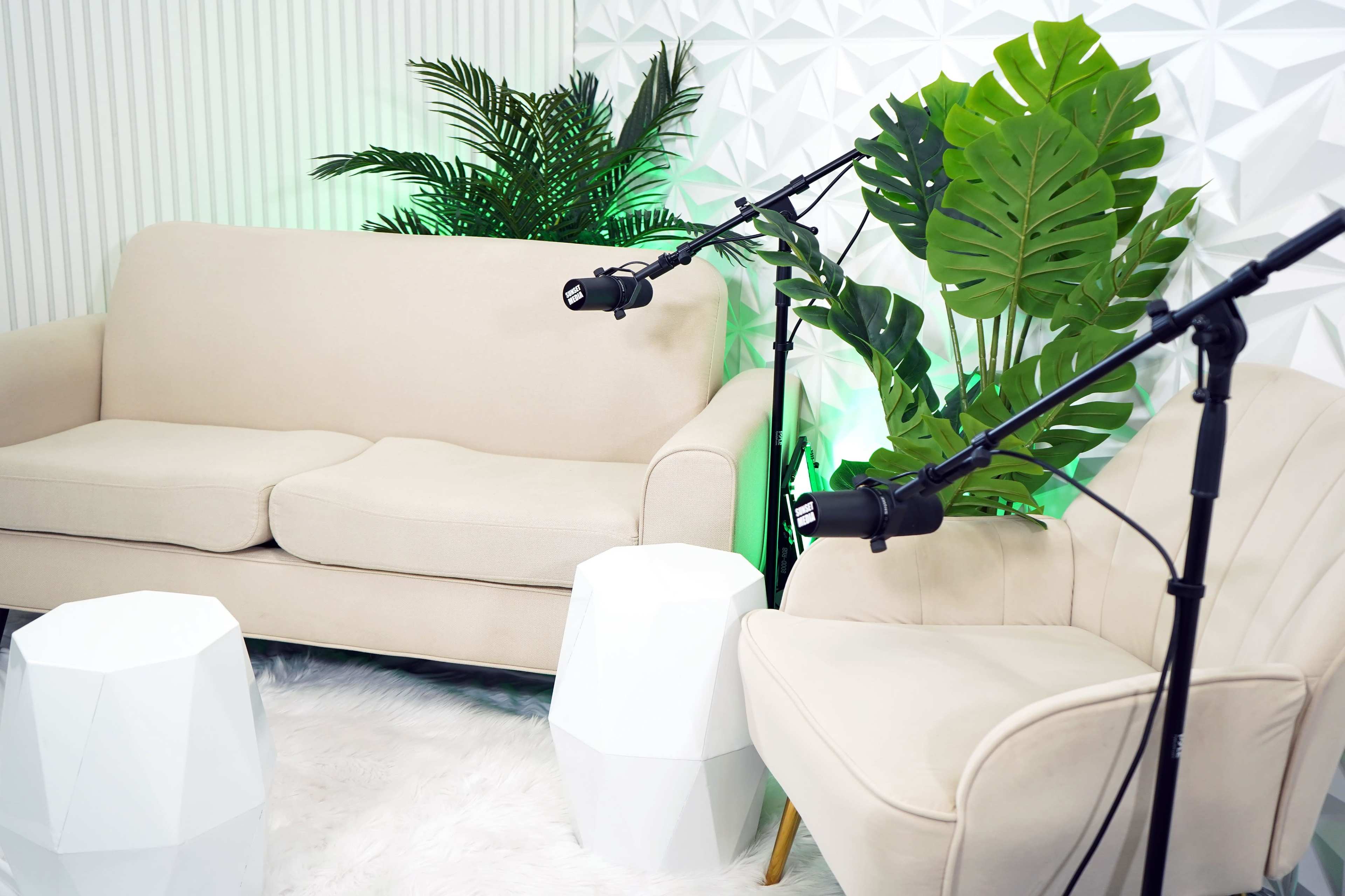 A modern seating area with a beige sofa, a white chair, and two adjustable lights, surrounded by large green plants against a textured wall.