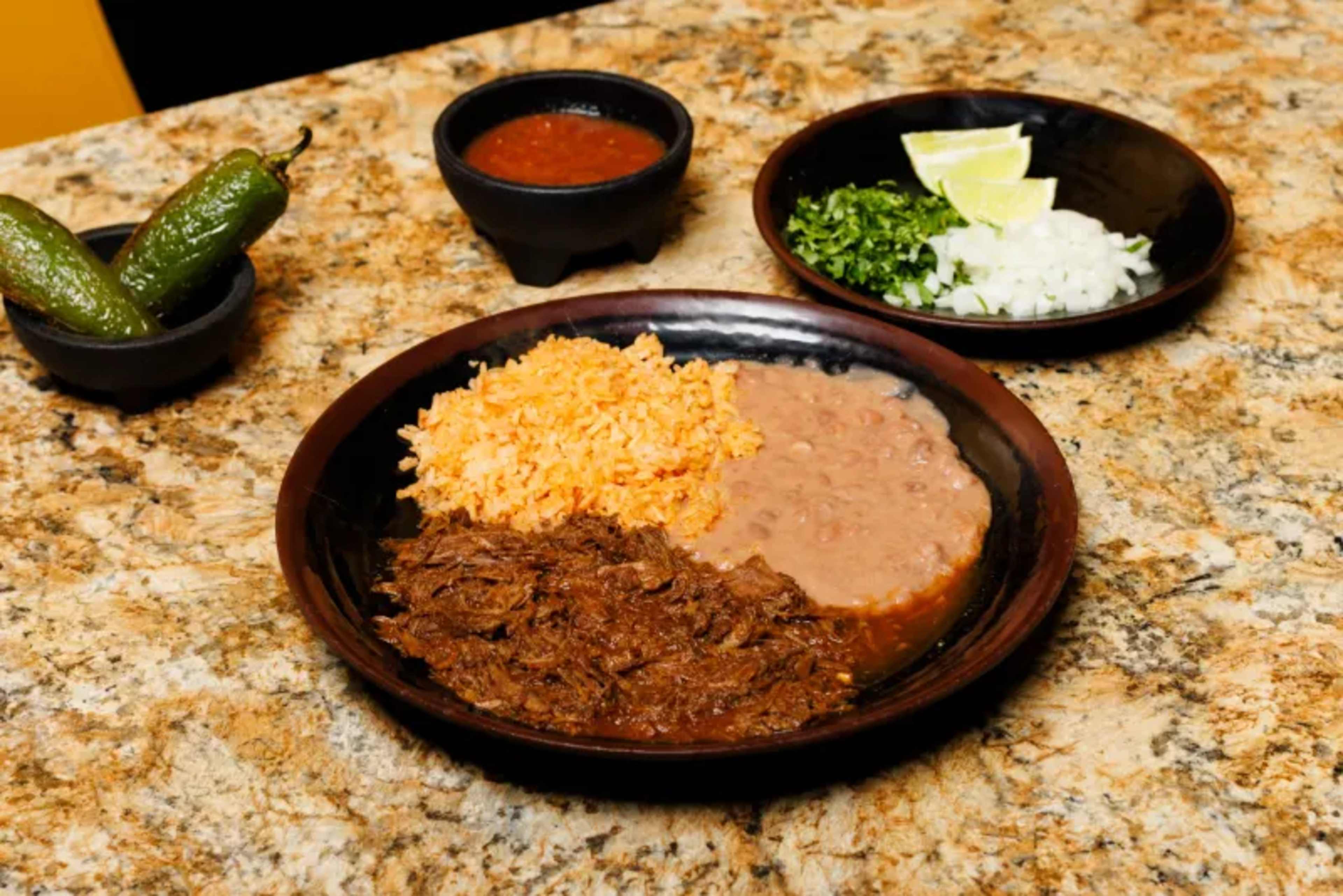 A plate of shredded beef with rice and refried beans is served alongside bowls of salsa, diced onions, chopped cilantro, lime wedges, and jalapeños on a granite countertop.