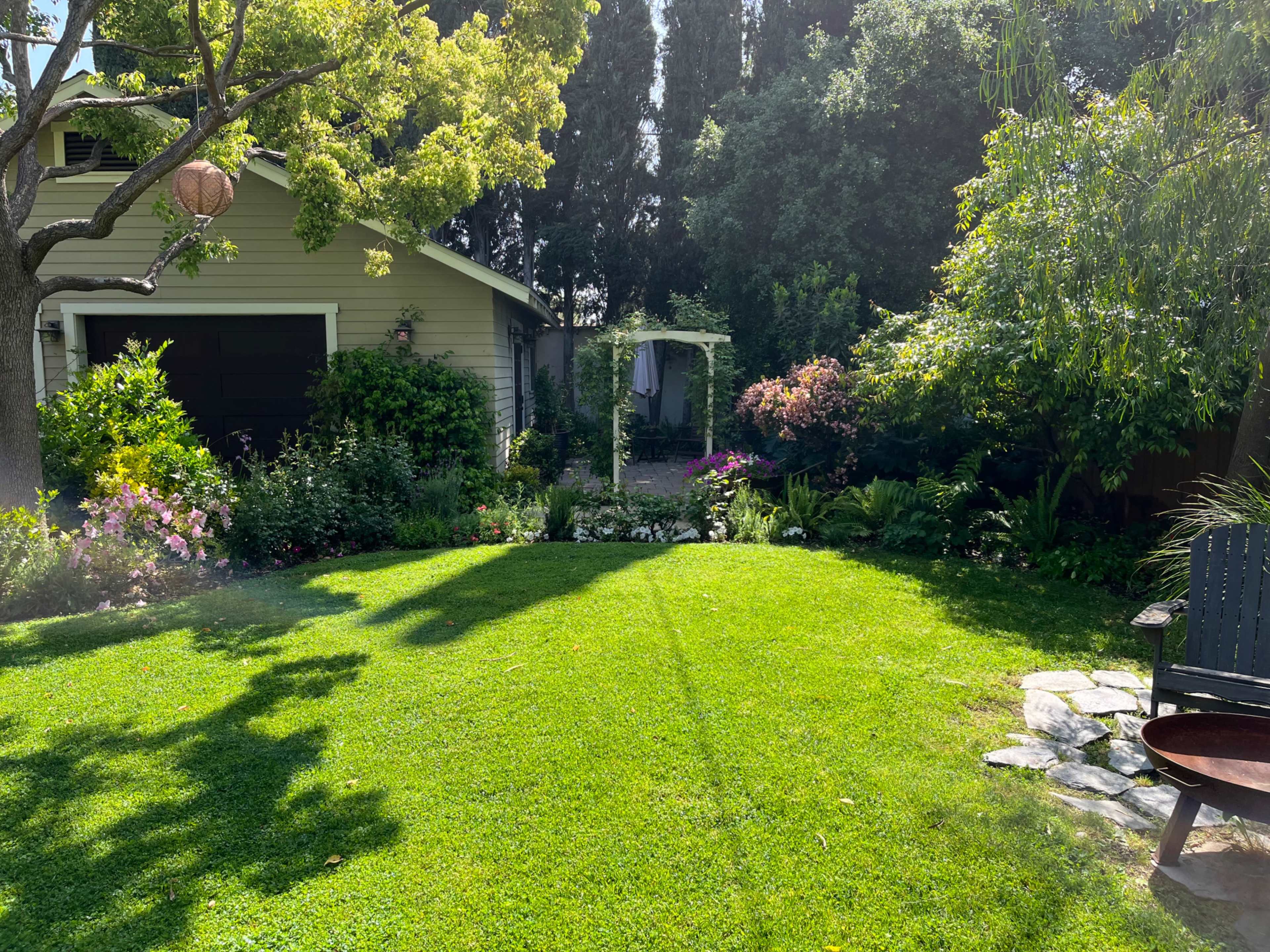 A well-maintained garden features a green lawn, flower beds, and a pathway leading to a white gazebo, with a house and trees in the background.