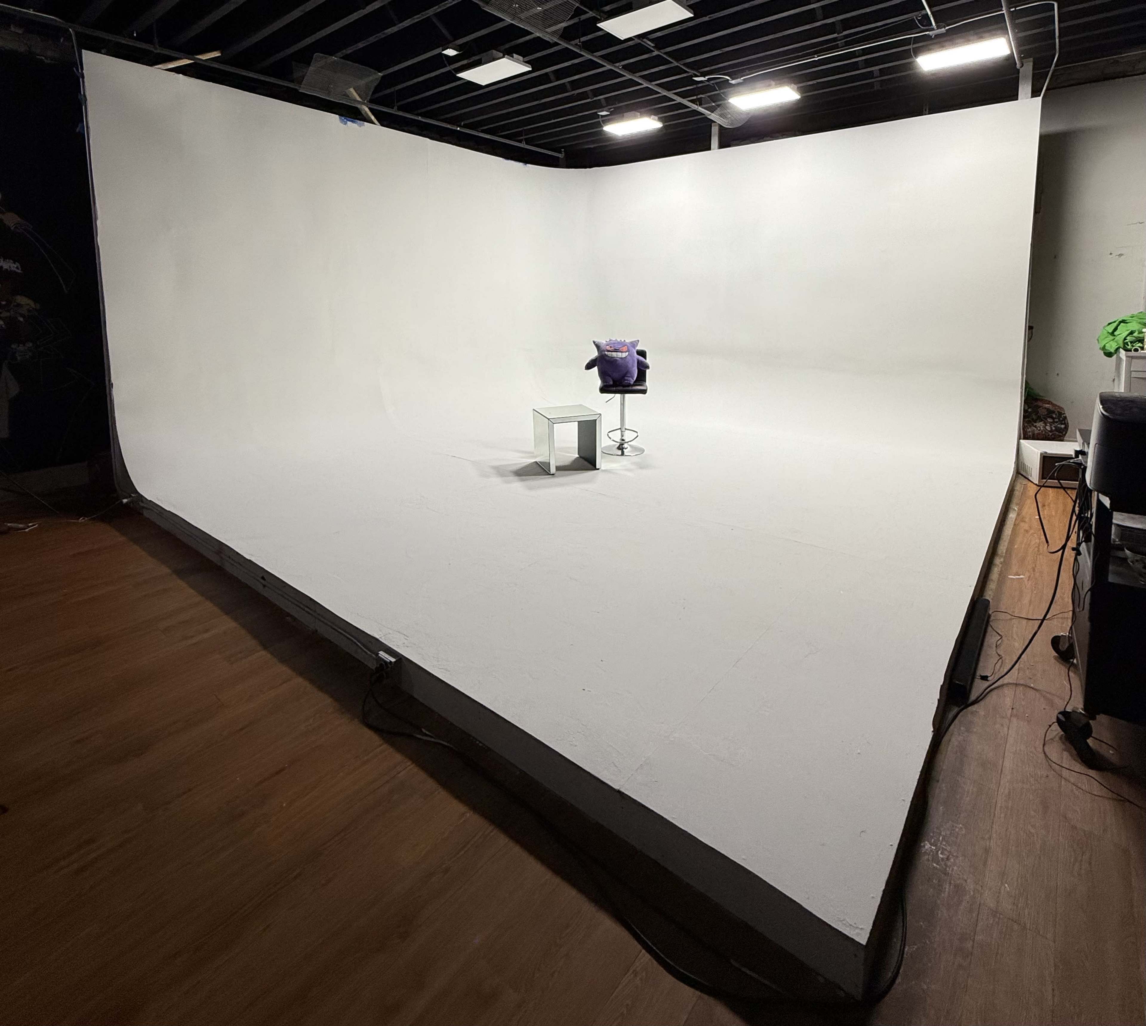 The image shows an empty studio with a large white backdrop, a stool, and a small table in the center, illuminated by overhead lights.