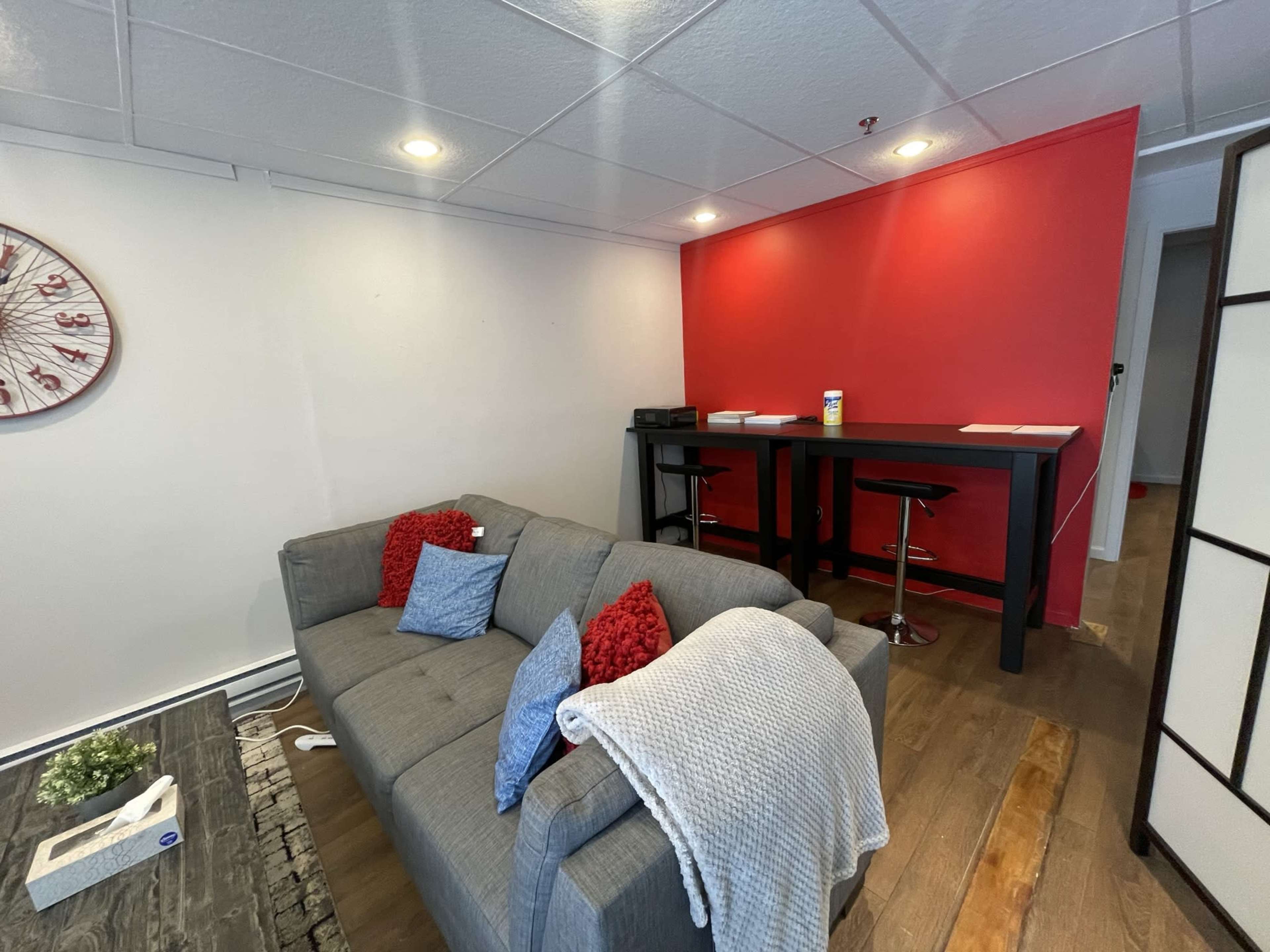 A gray couch with red pillows sits in a room next to a black bar table with two stools against a red wall.