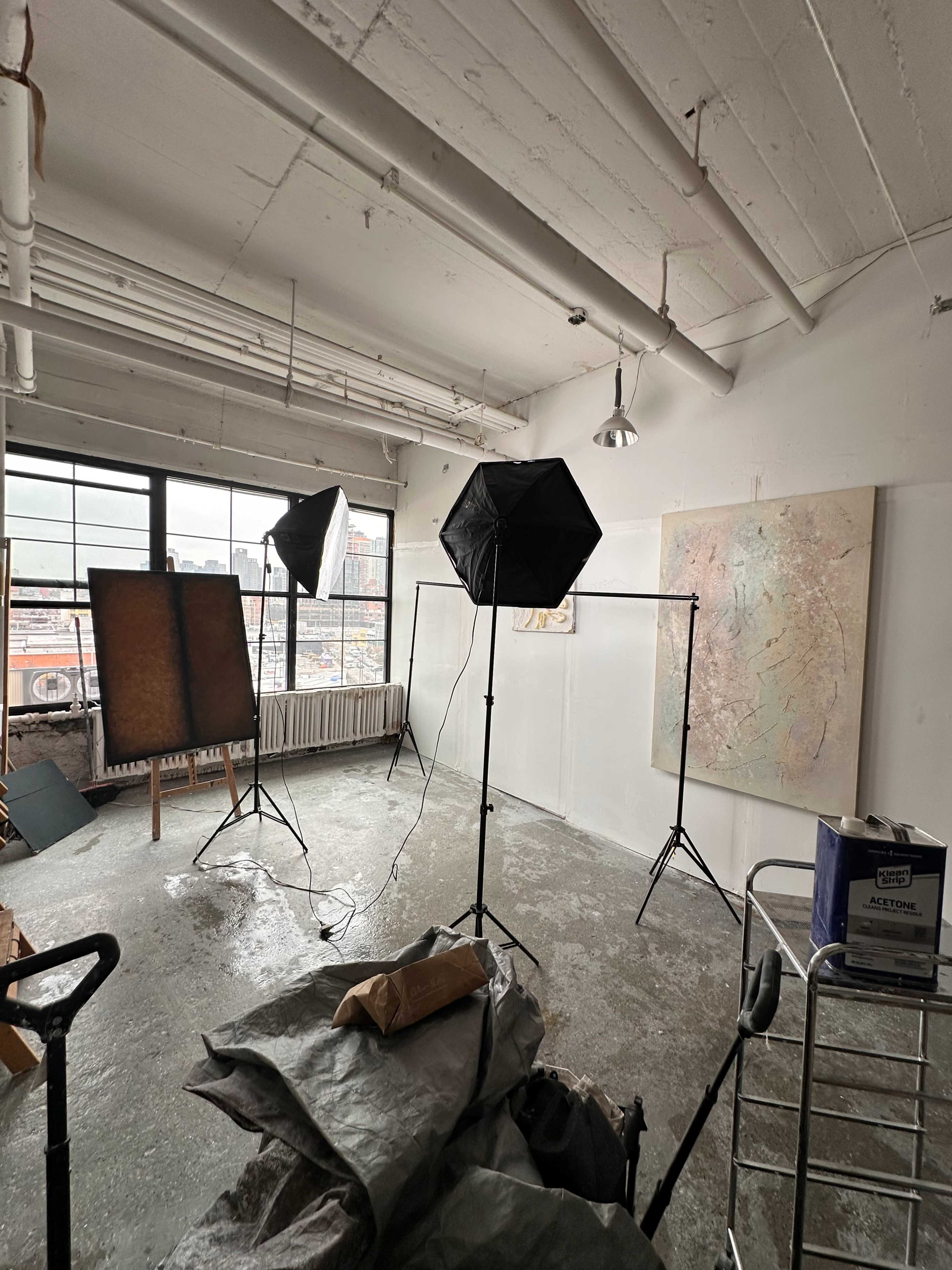 The image shows an empty art studio with two photography lights, a large window, and several art pieces displayed, including an abstract painting on the wall.