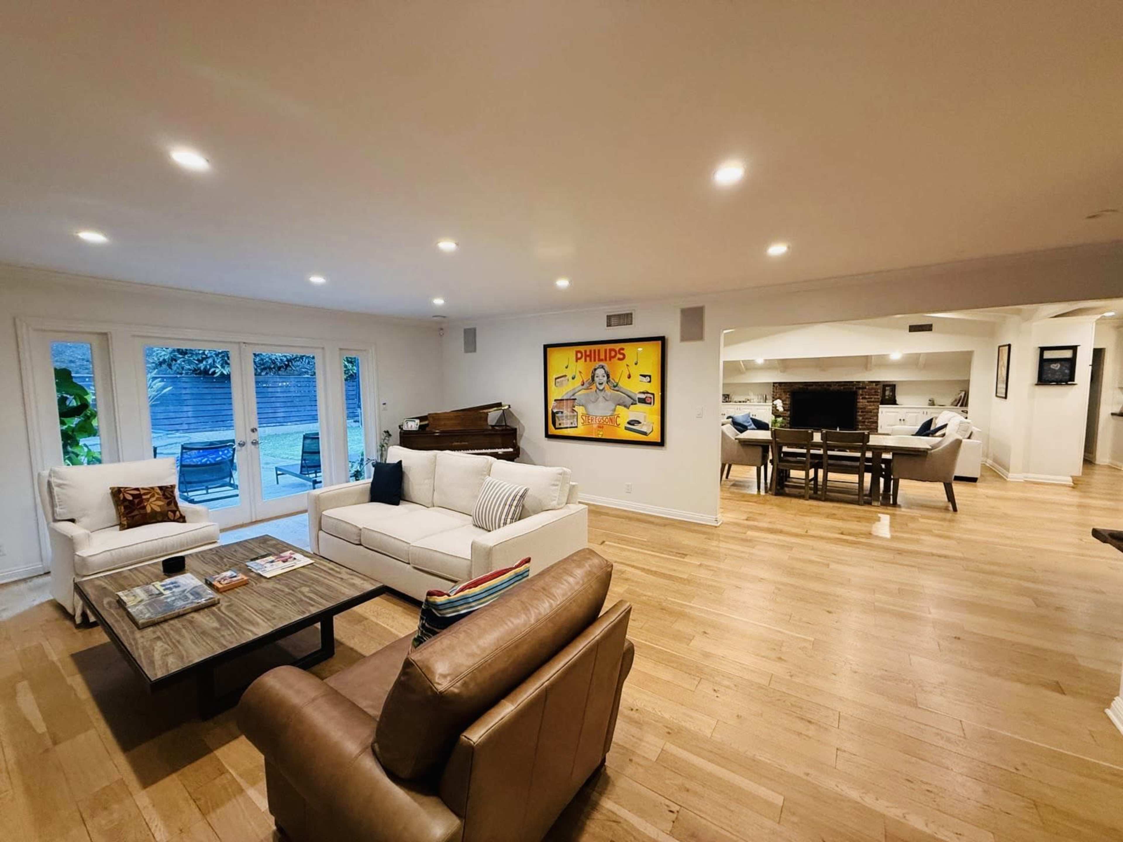 The image shows a spacious living room with a white sofa, a wooden coffee table, a leather armchair, and a brightly colored artwork on the wall, opening into an adjacent dining area.