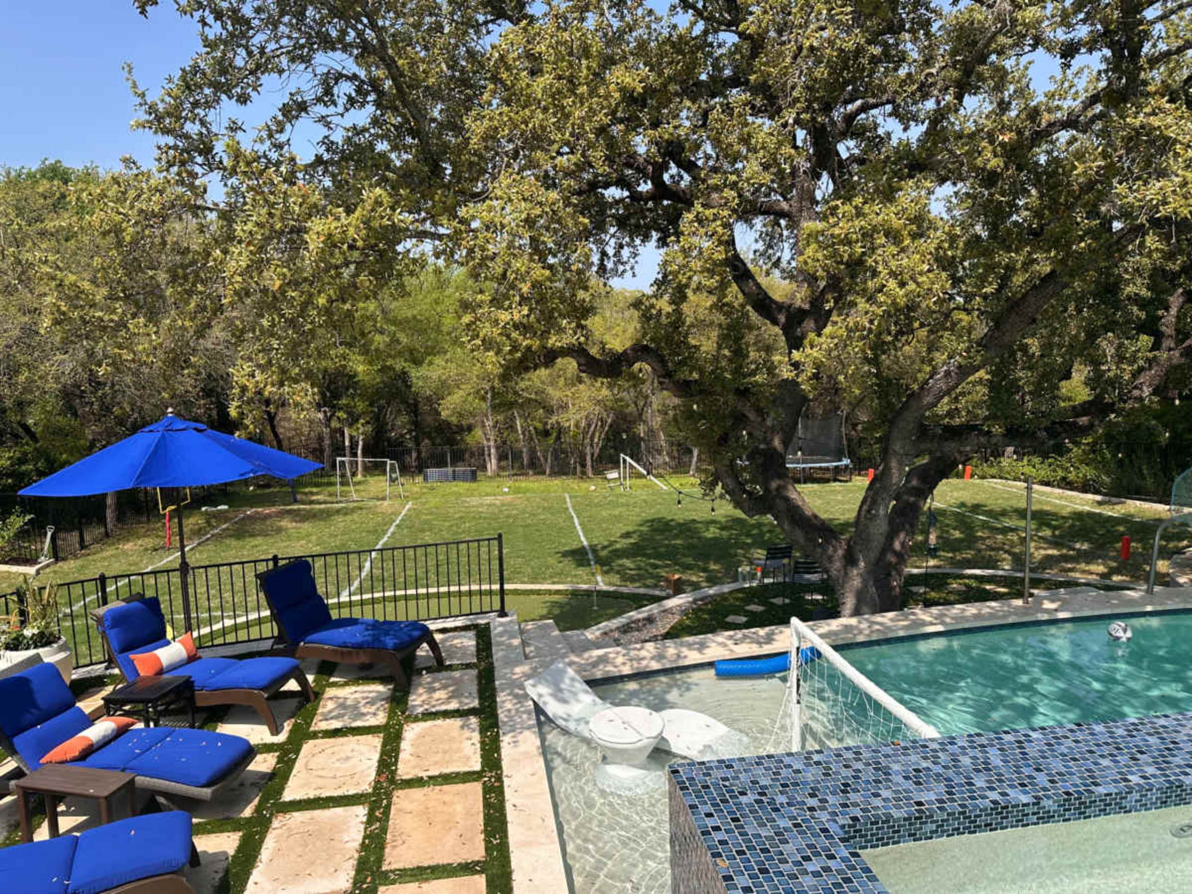 A backyard features a swimming pool, lounge chairs, and a large grassy area with soccer goals in the distance.
