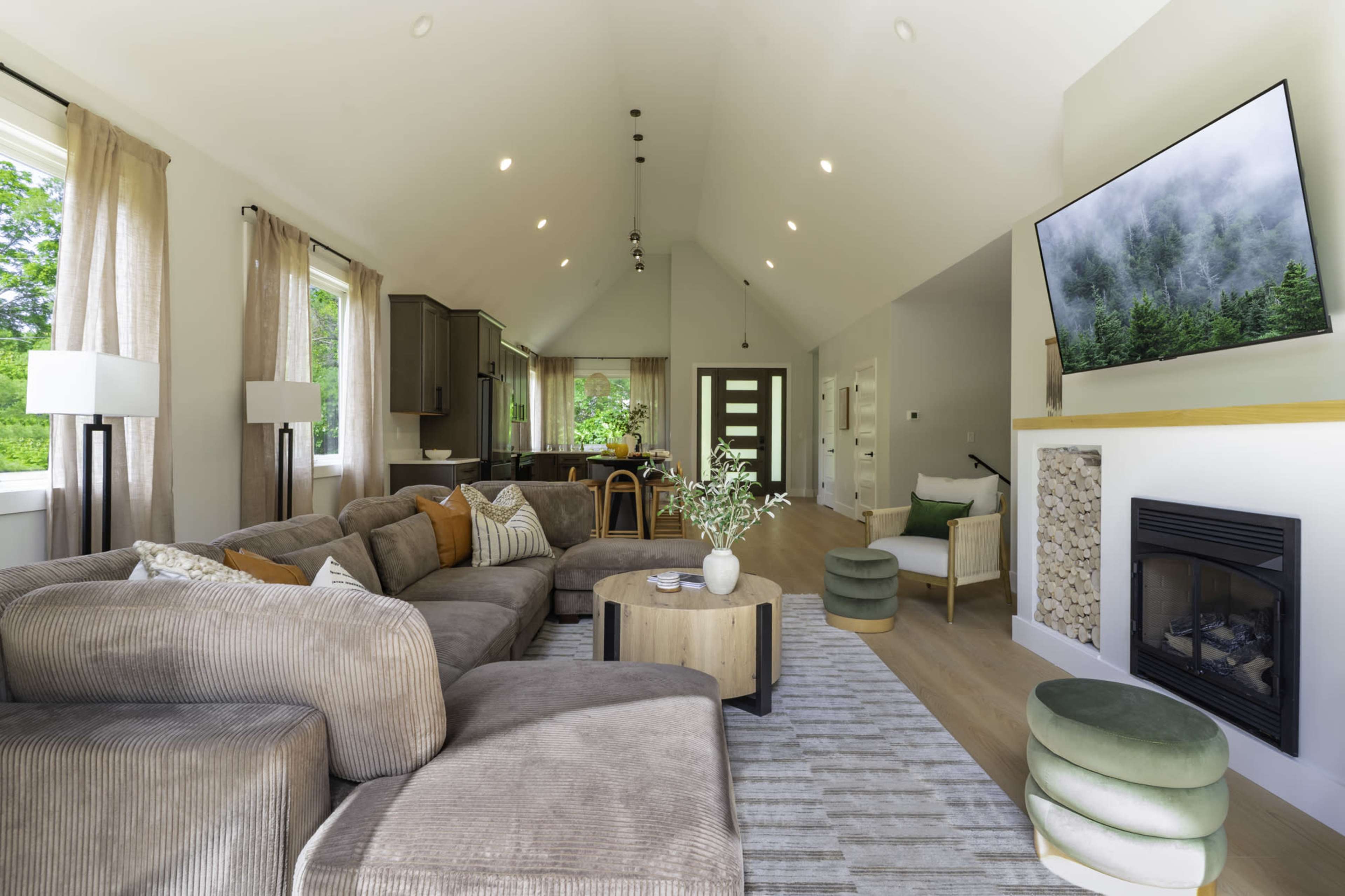 The image shows a modern living room with a large sectional sofa, a coffee table, a mounted television, and a fireplace, all set against a backdrop of large windows allowing natural light.