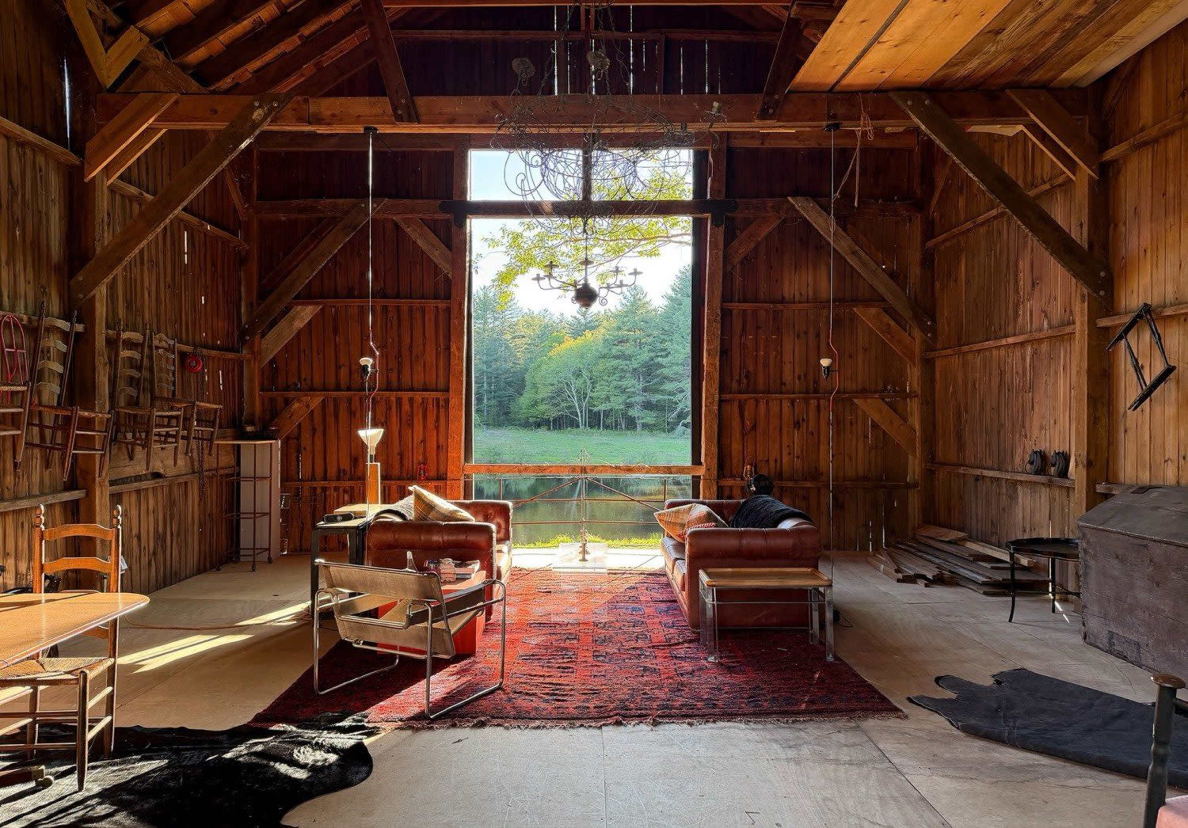 An interior of a rustic wooden barn features seating areas with a view of a green landscape through a large opening.