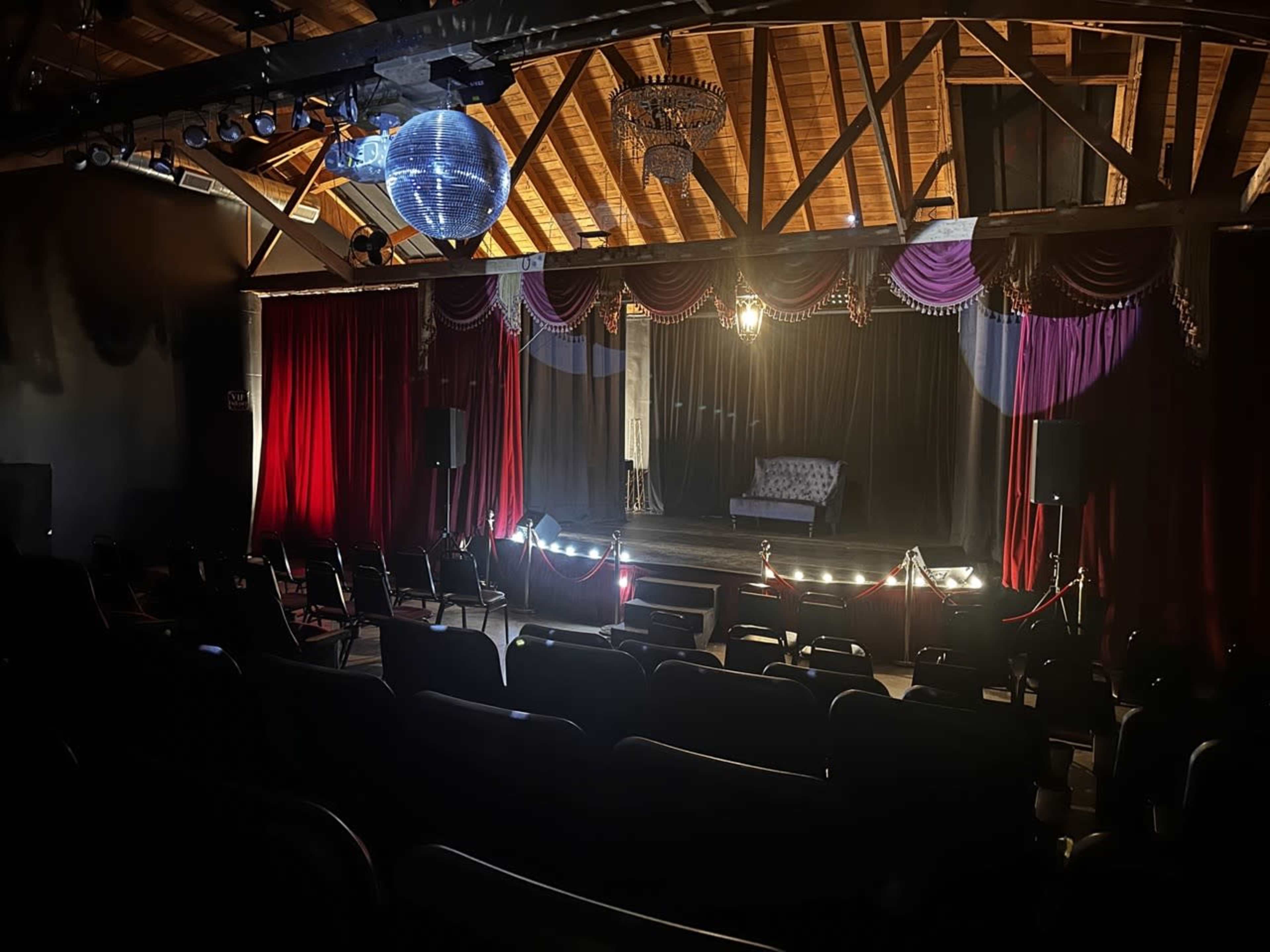 A dimly lit theater features a stage with a single sofa and decorative curtains, surrounded by rows of empty chairs.