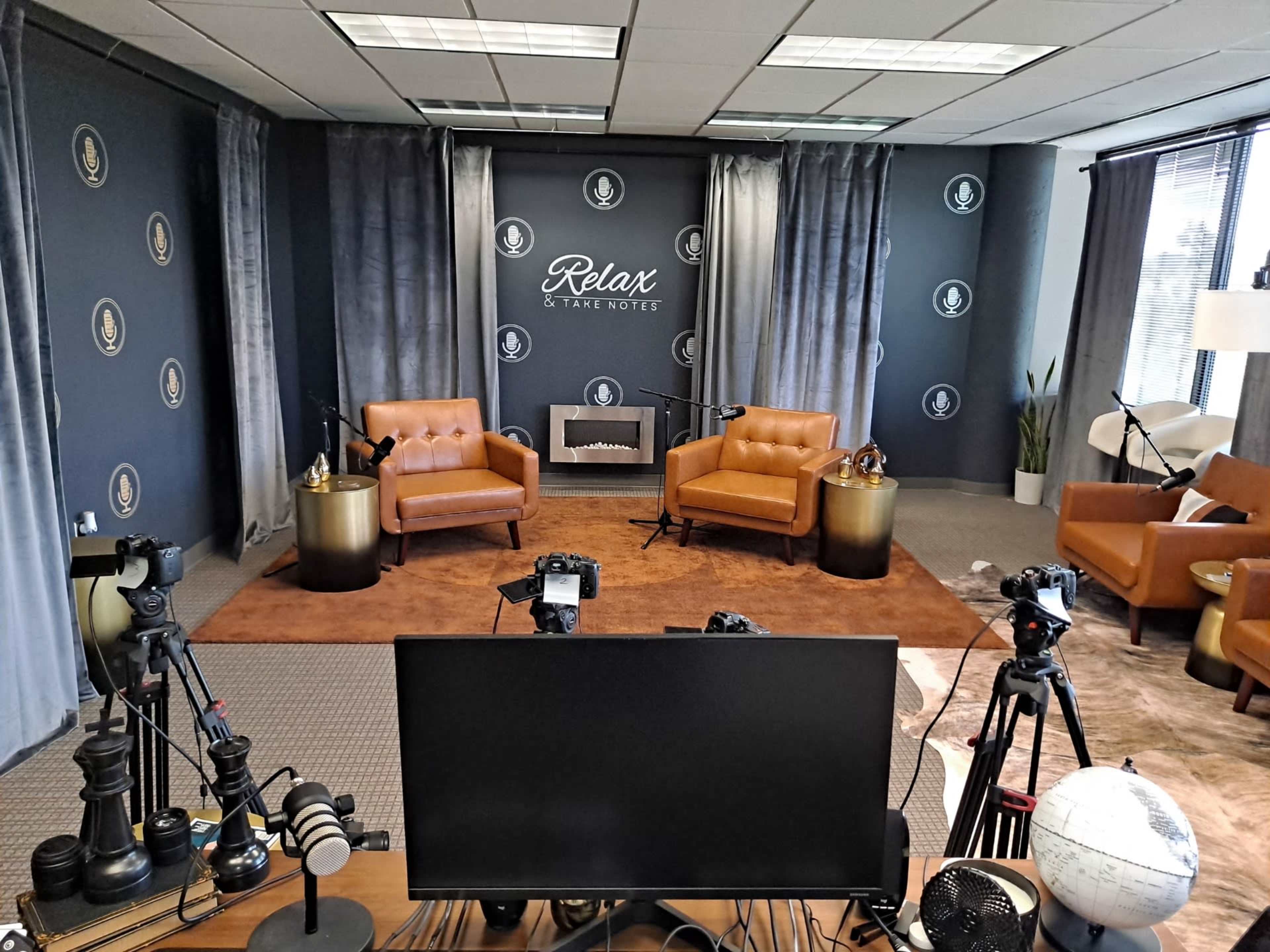 A recording studio with two brown leather chairs, a coffee table, various cameras, and a backdrop featuring the phrase "Relax & Take Notes."