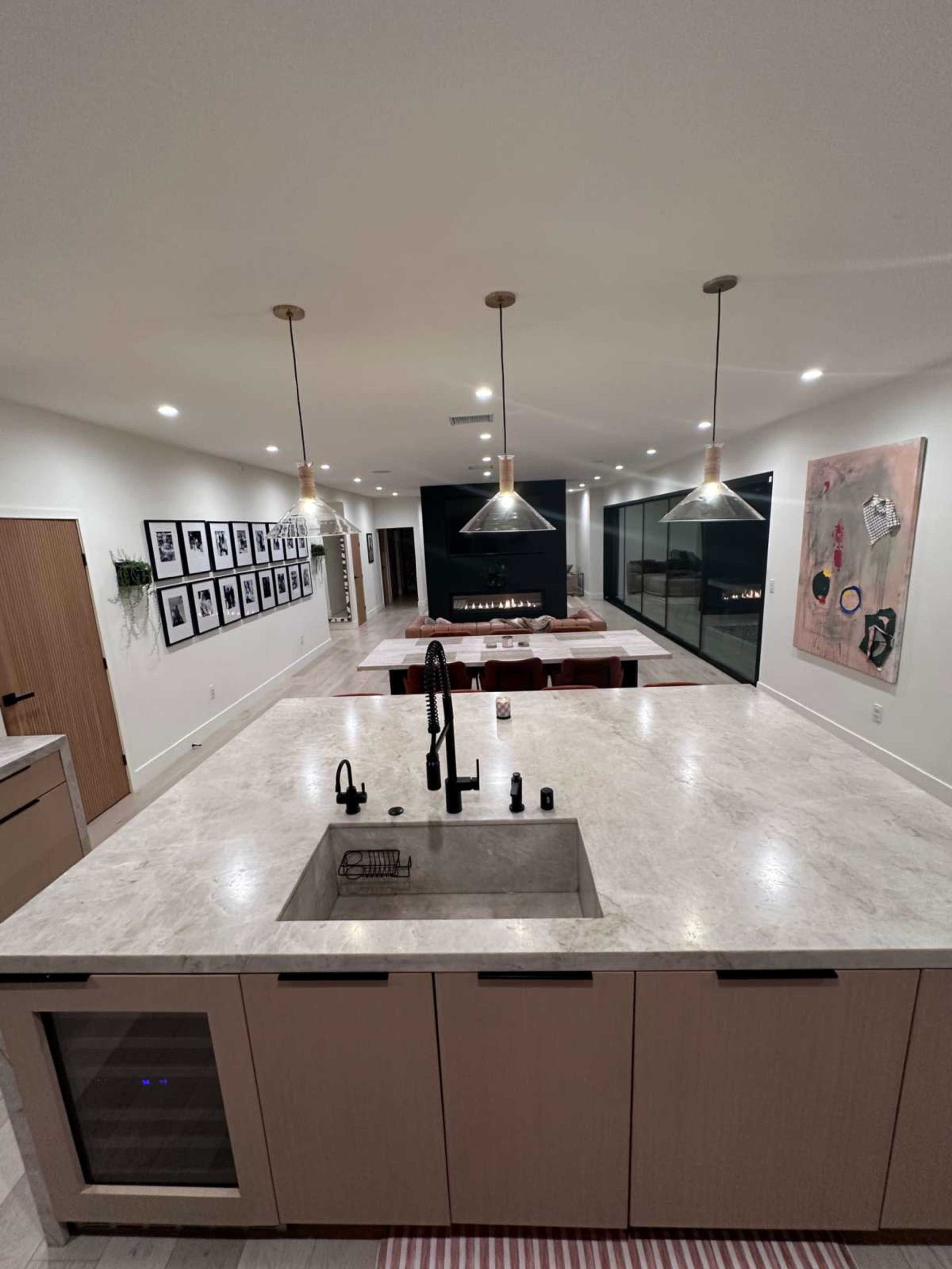 The image shows a modern kitchen with a marble counter and sink, overlooking an open living area featuring a fireplace and a wall adorned with framed pictures.