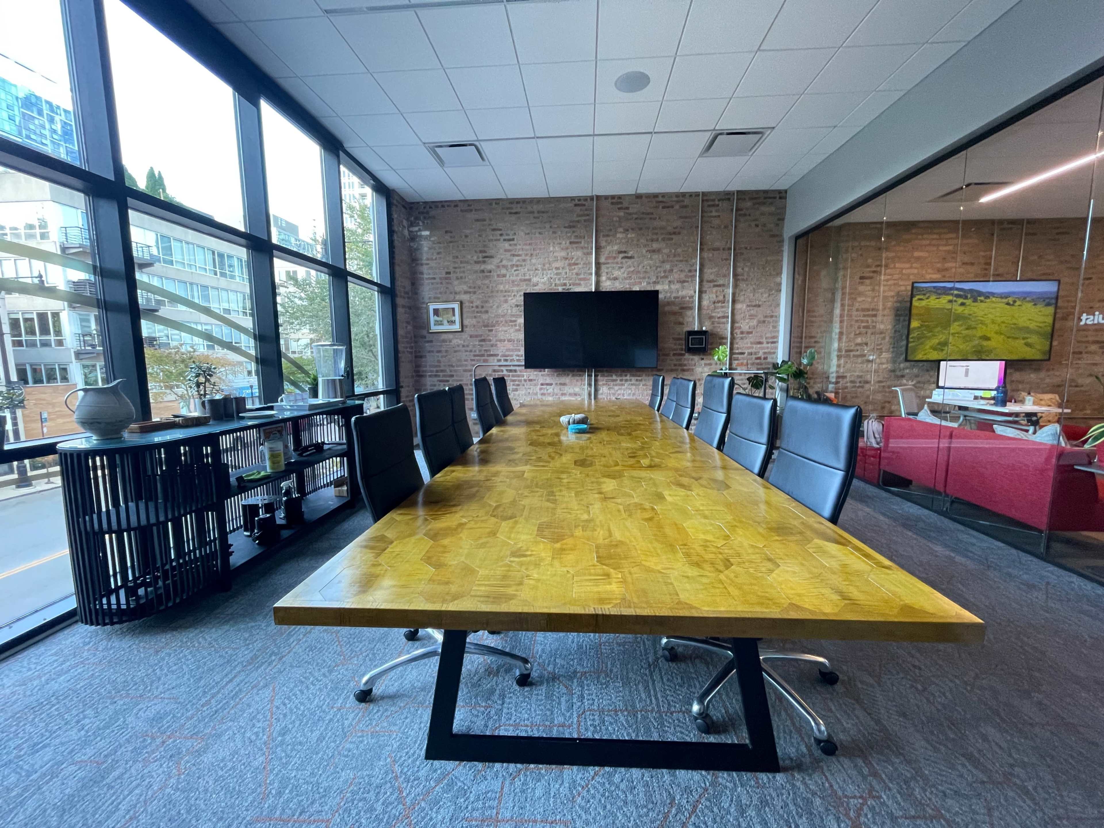 The image shows a modern conference room featuring a long wooden table surrounded by black office chairs, with large windows allowing natural light and a view of the outside.