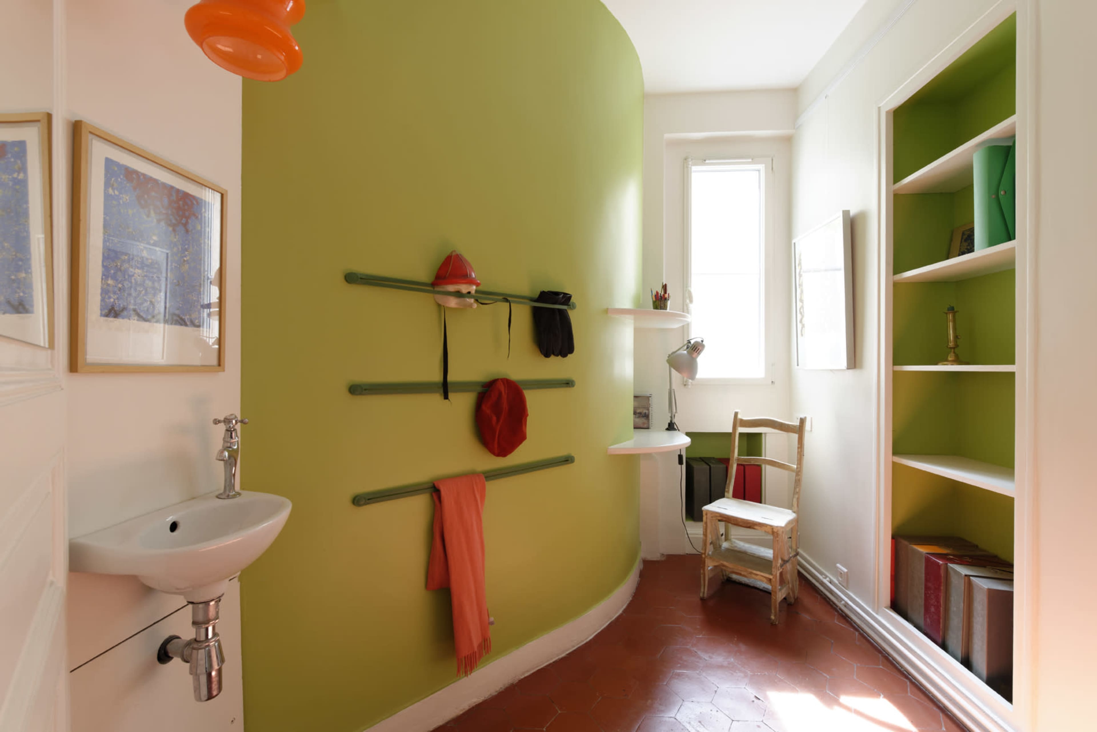 A small room with a bright green curved wall, a sink, shelves on one side, and a wooden chair near a window.