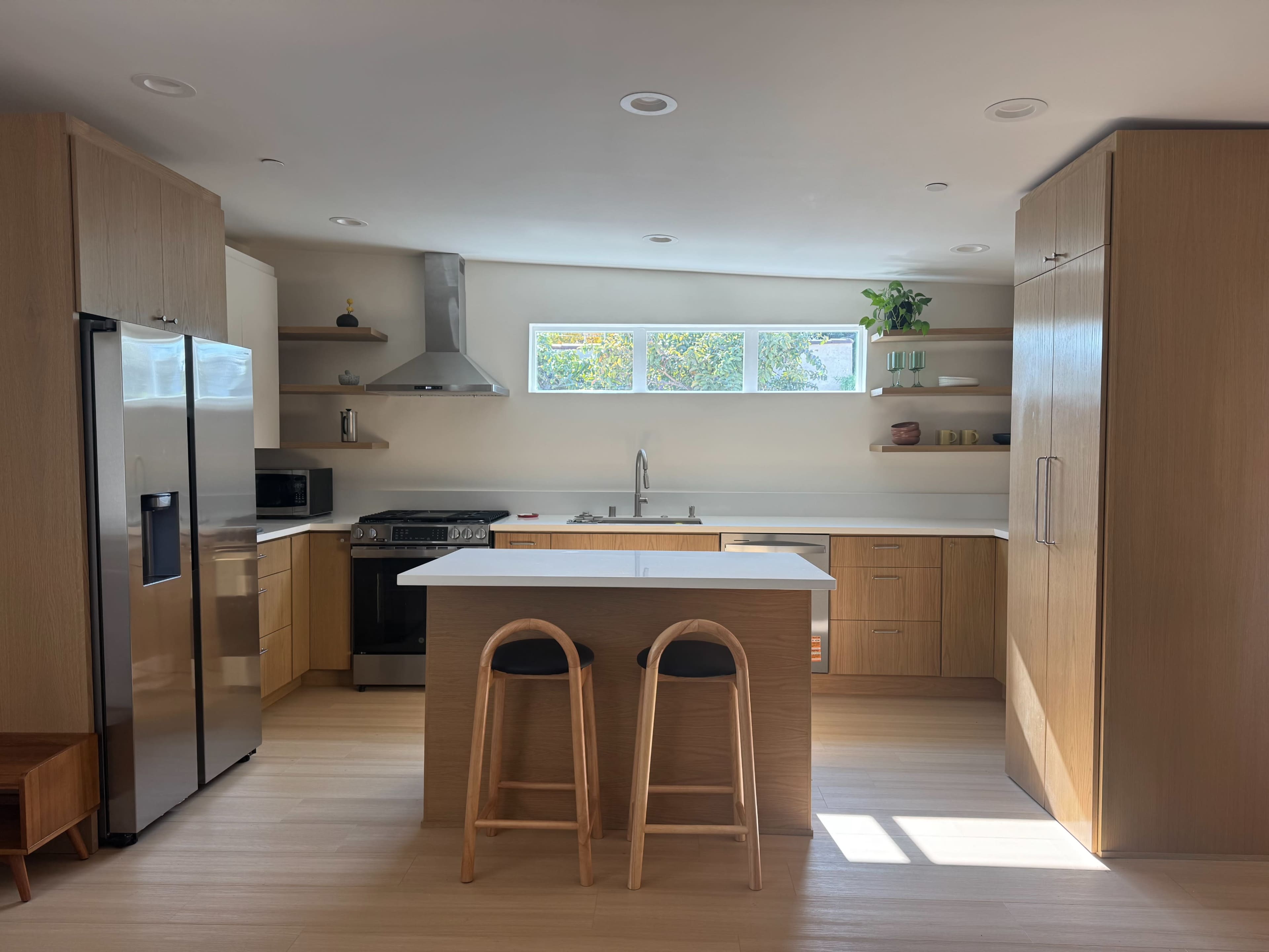 The image shows a modern kitchen featuring wooden cabinetry, stainless steel appliances, and a central island with two wooden stools.