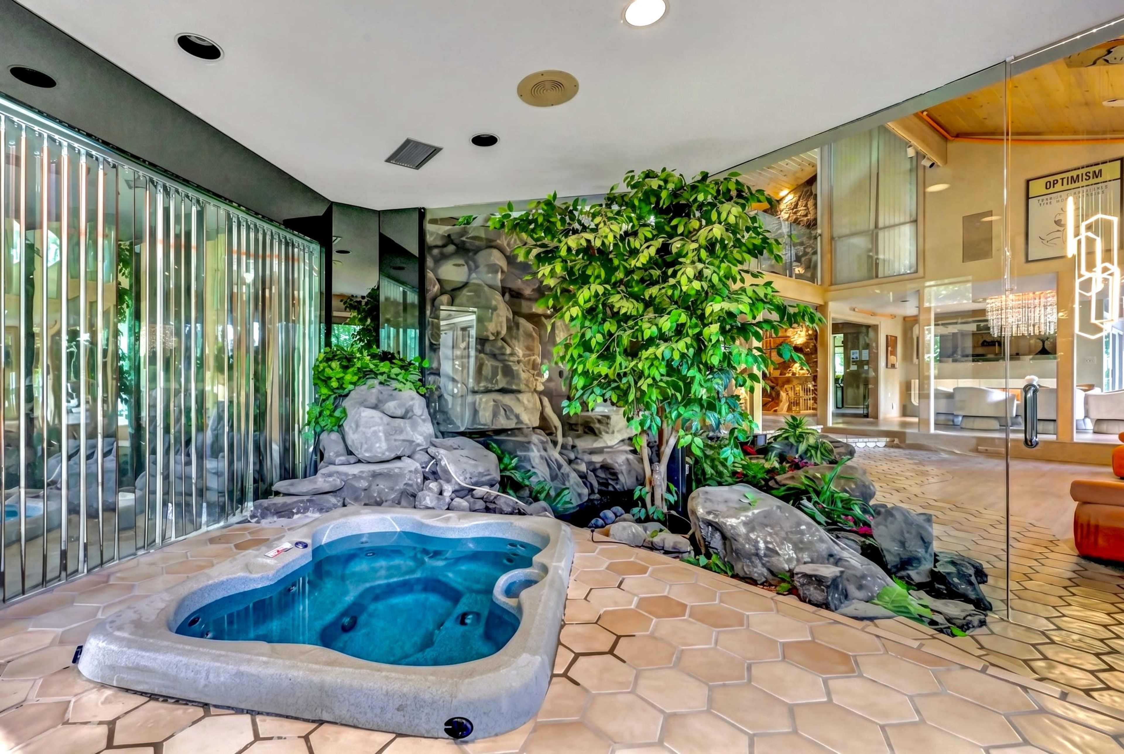 The image shows a sunlit indoor space featuring a small hot tub surrounded by rocks and greenery, with large glass walls providing a view of a modern living area.
