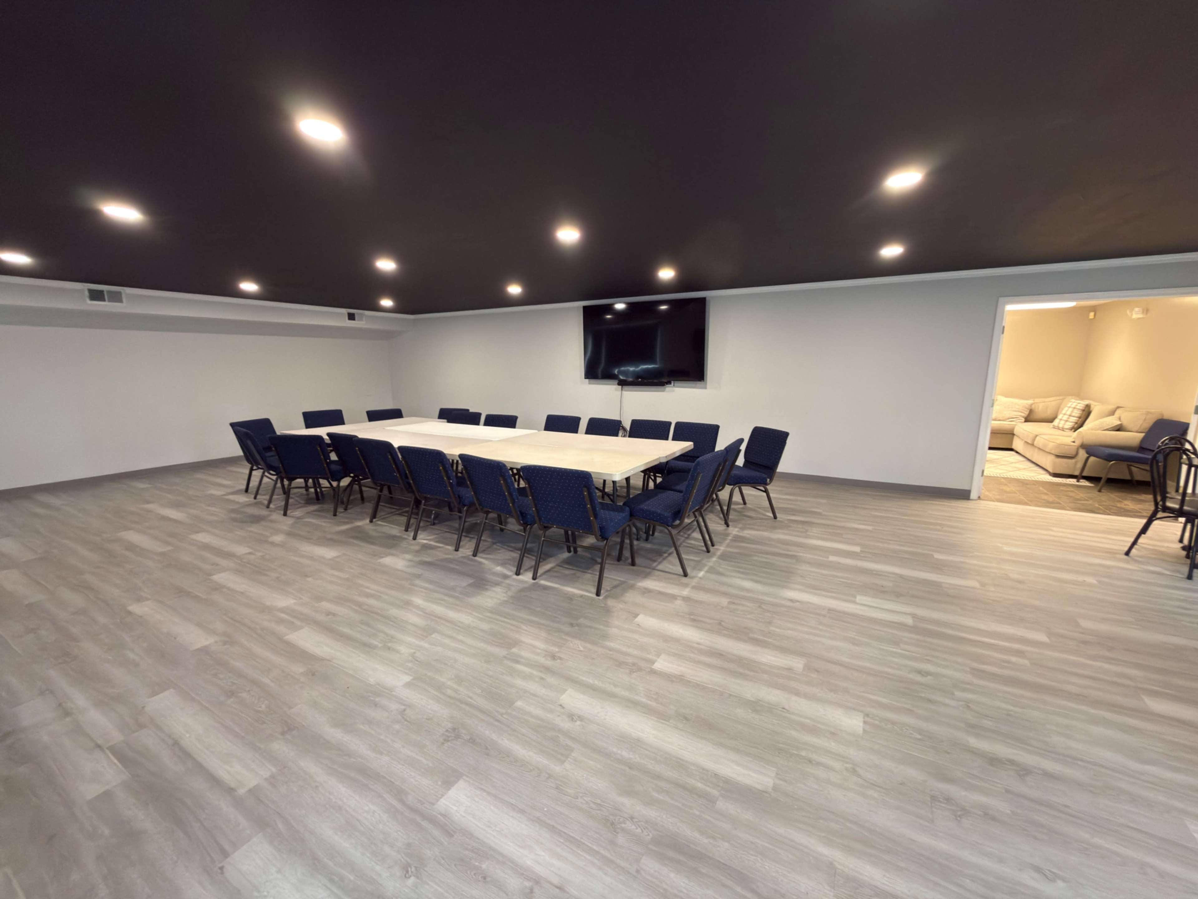 A large conference room features a long rectangular table surrounded by blue chairs, with a television mounted on the wall and a doorway leading to a seating area.
