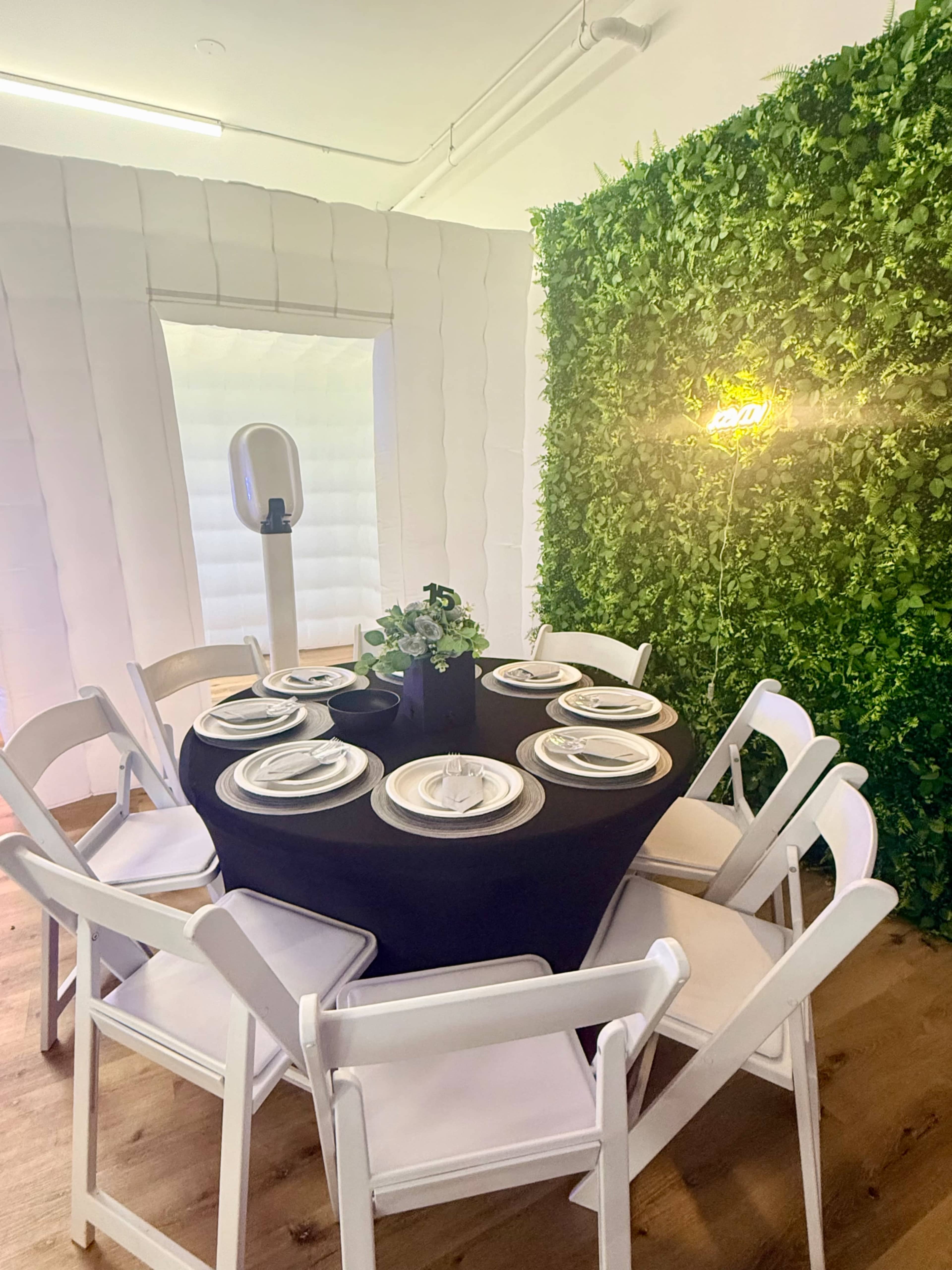 A round table with a black cloth is set for a meal with plates and silverware, surrounded by white chairs in a room featuring a green wall of foliage.