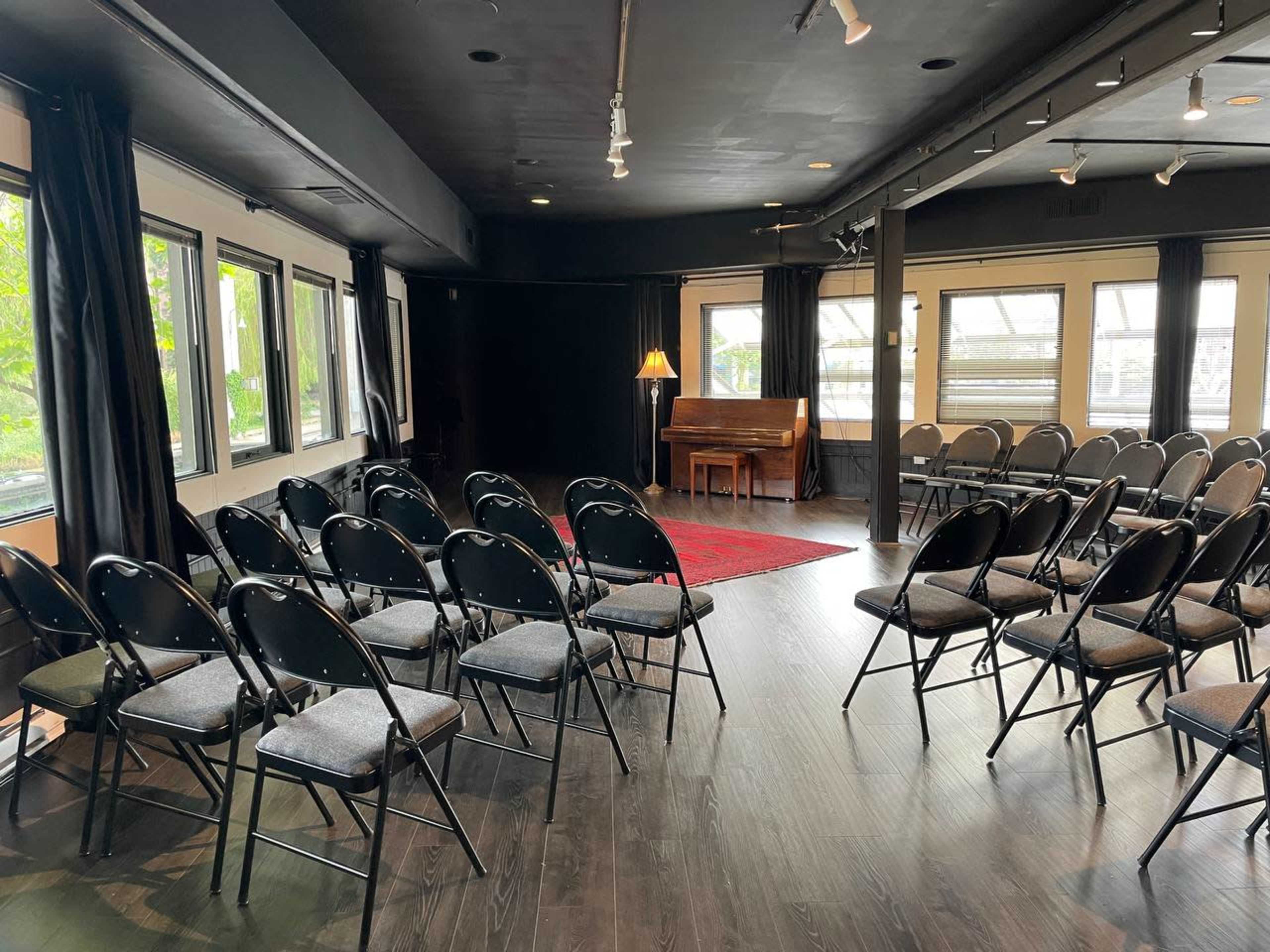 A small indoor performance space features rows of folding chairs facing a red rug and a piano, with large windows on one side.