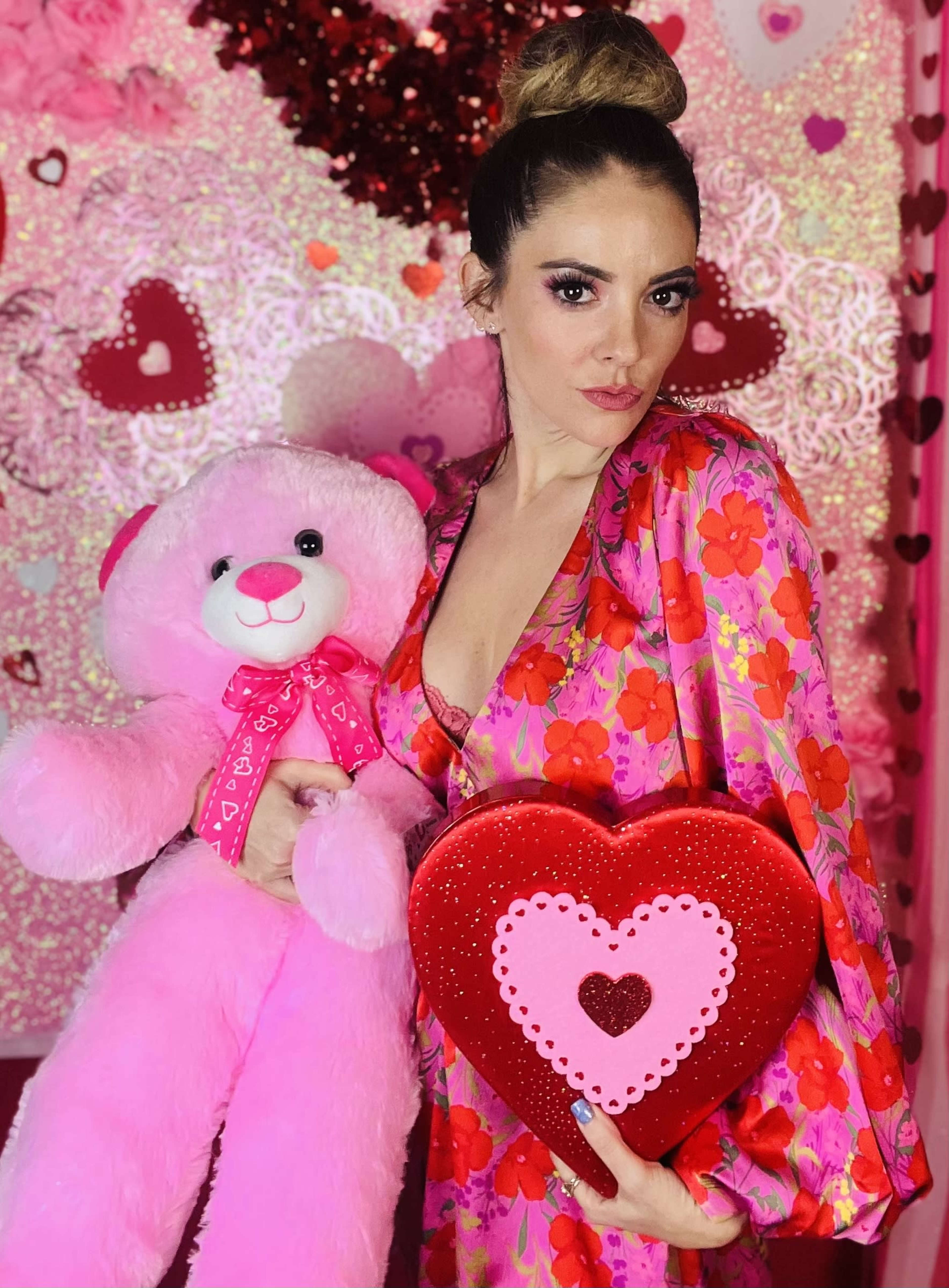 A woman poses with a large pink teddy bear and a red heart-shaped box in front of a pink and heart-themed background.