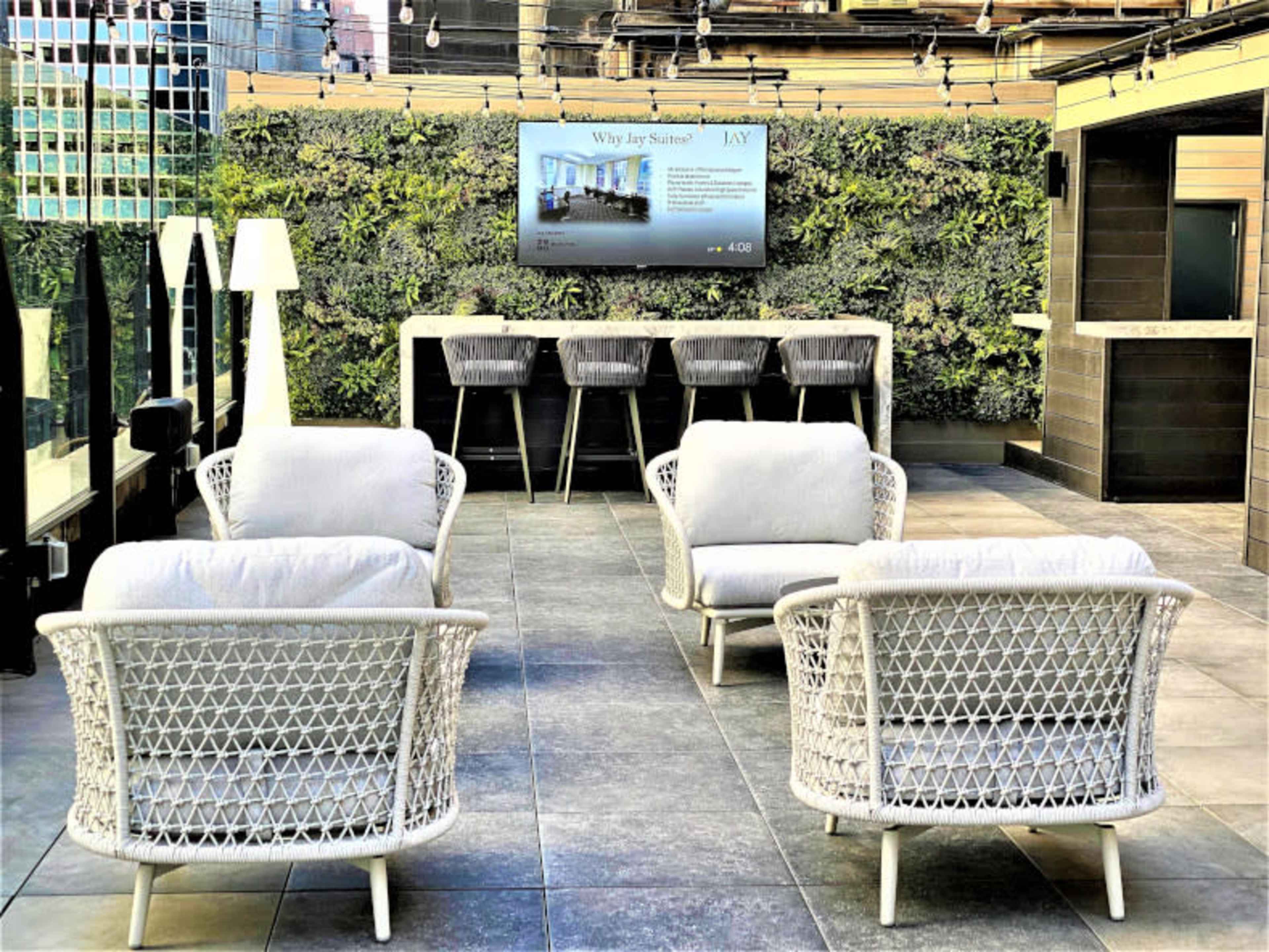 The image shows a modern rooftop lounge area with four white wicker chairs arranged around a small table, a wall of greenery behind, and a digital display mounted on the wall.