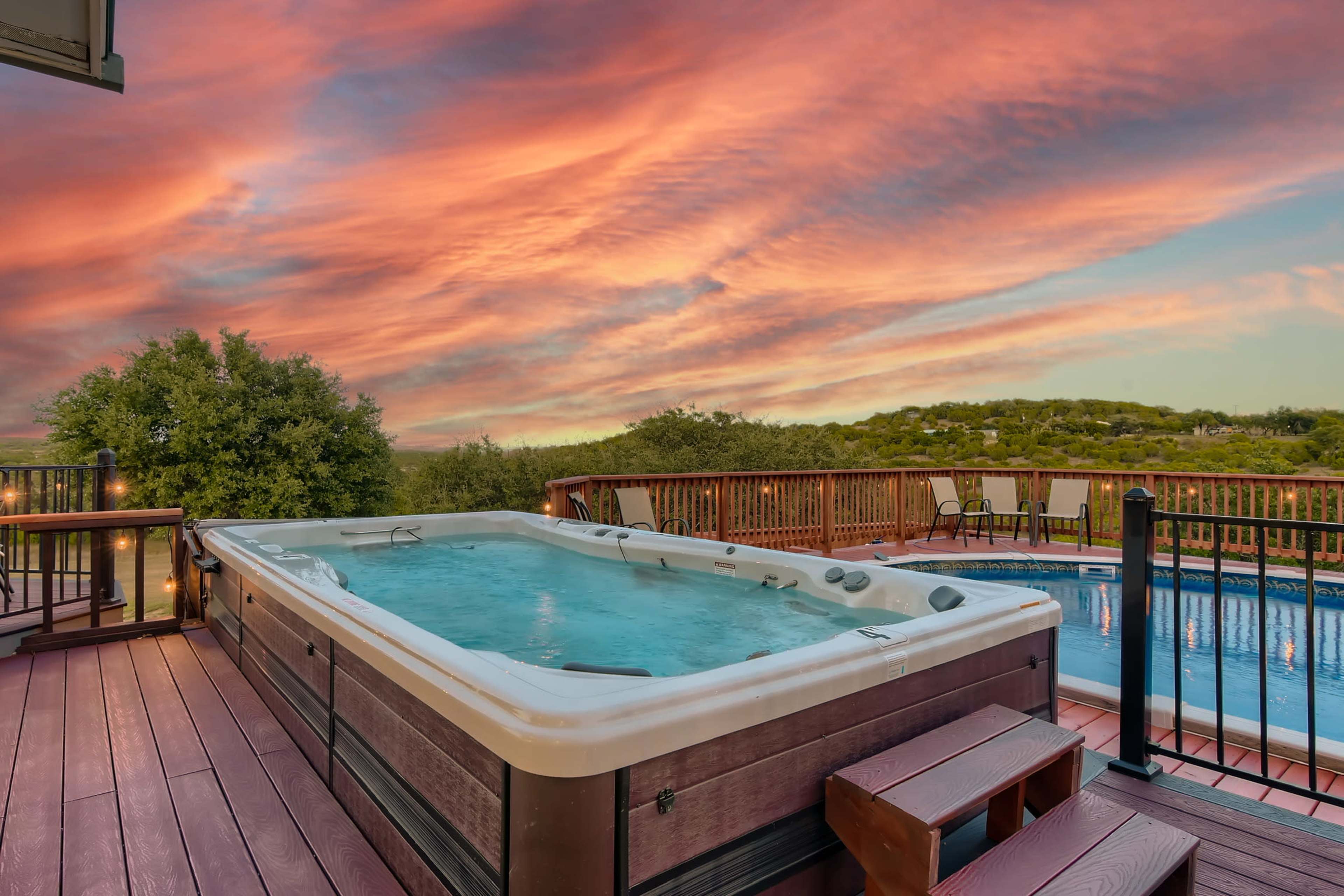 A hot tub is positioned on a wooden deck overlooking a pool, with a colorful sunset sky in the background.