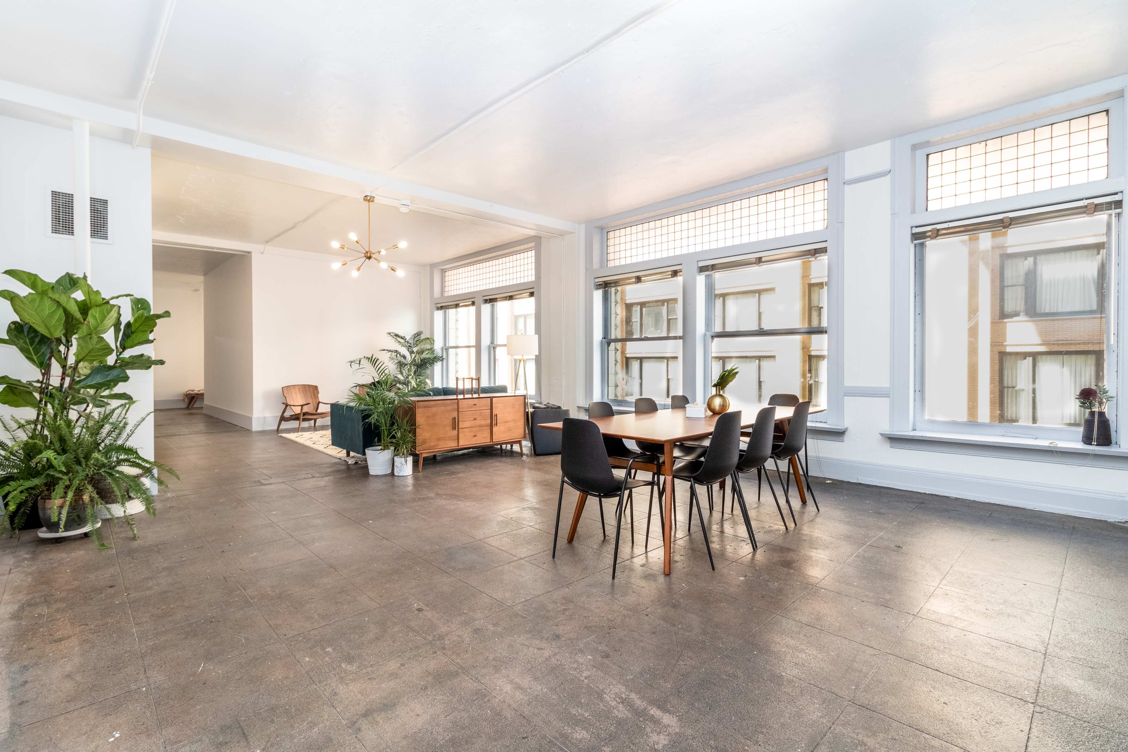 A spacious room features a dining table surrounded by black chairs, large windows, and various indoor plants.