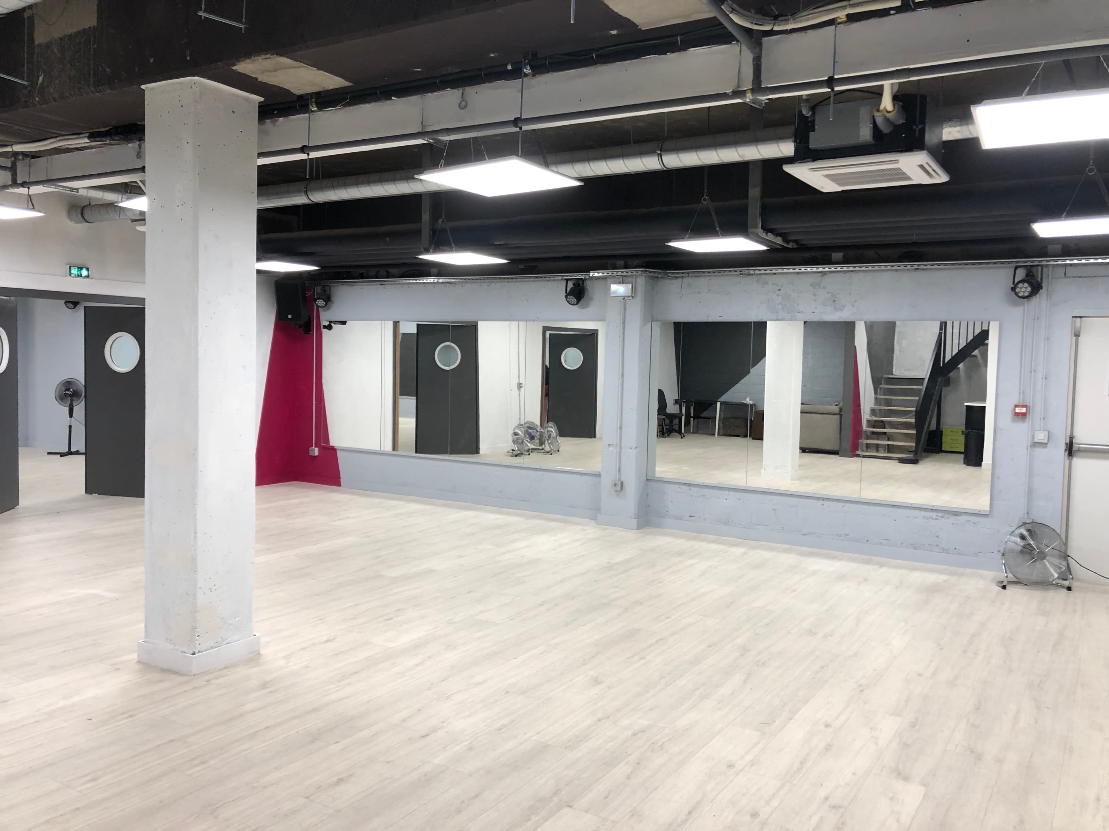 A spacious, empty studio with two large mirrors, gray walls, and separate doors, along with a staircase in the background.