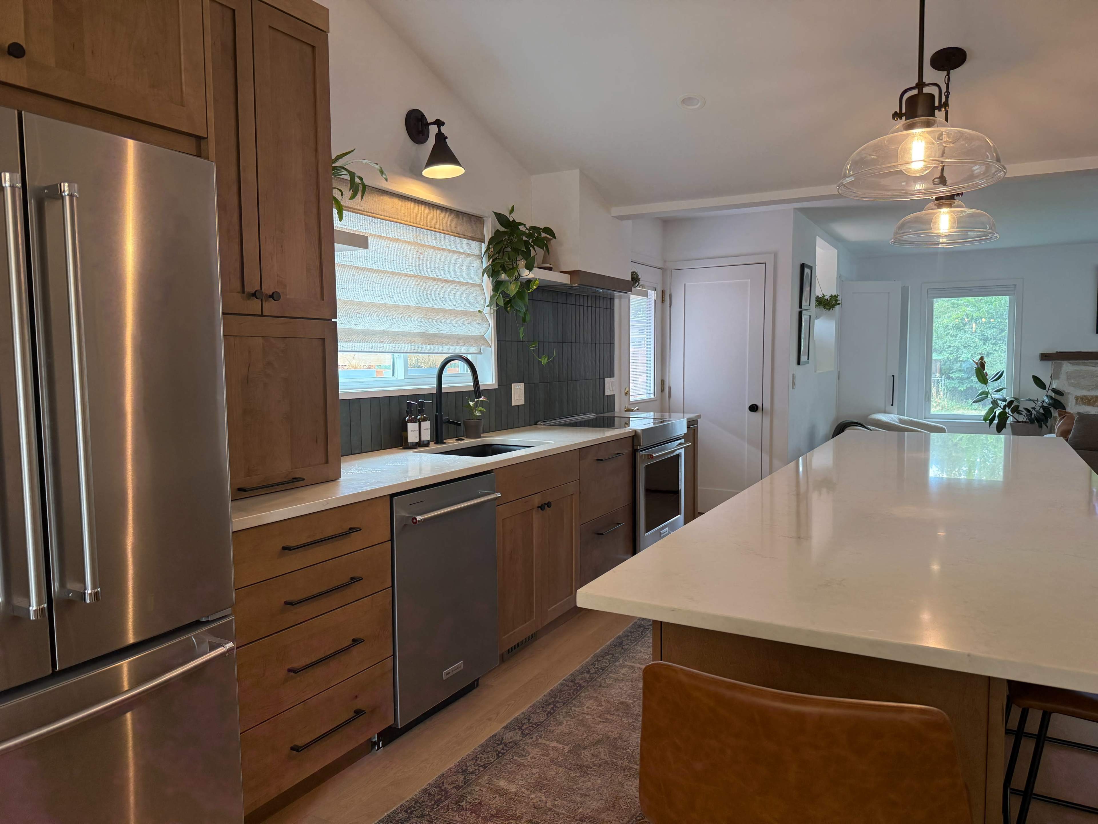 The image shows a modern kitchen with wooden cabinets, stainless steel appliances, a large island with seating, and plants along the countertop.